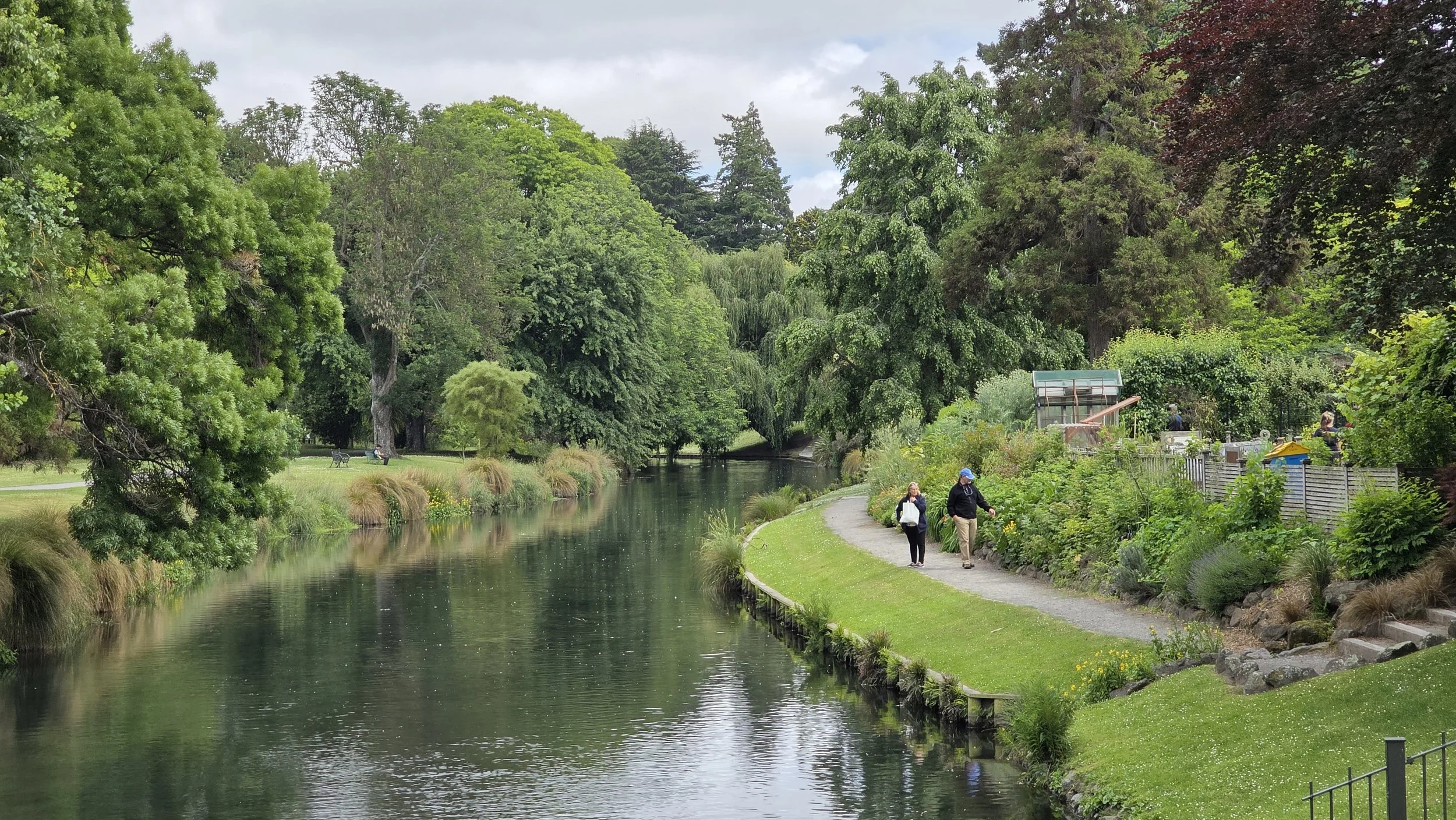 A peaceful park scene with a waterway and walking path, surrounded by lush green trees and plants, with a few people strolling along the path.