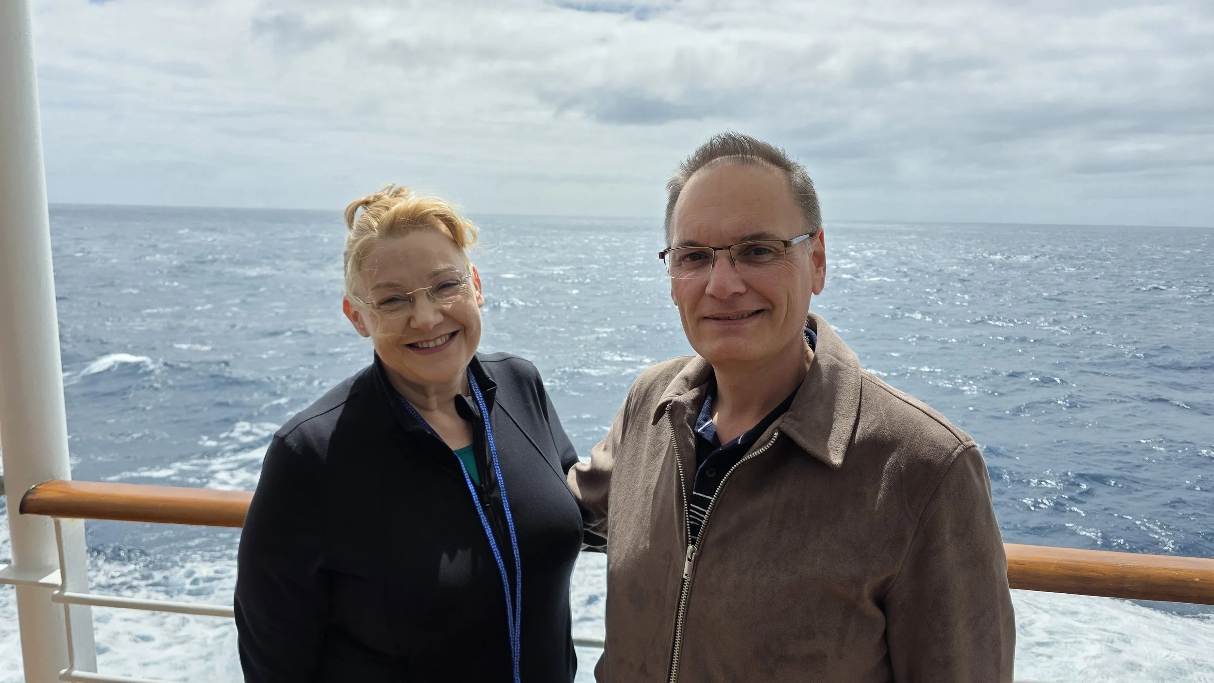 A smiling woman and man standing on the deck of a boat with the ocean and cloudy sky in the background.