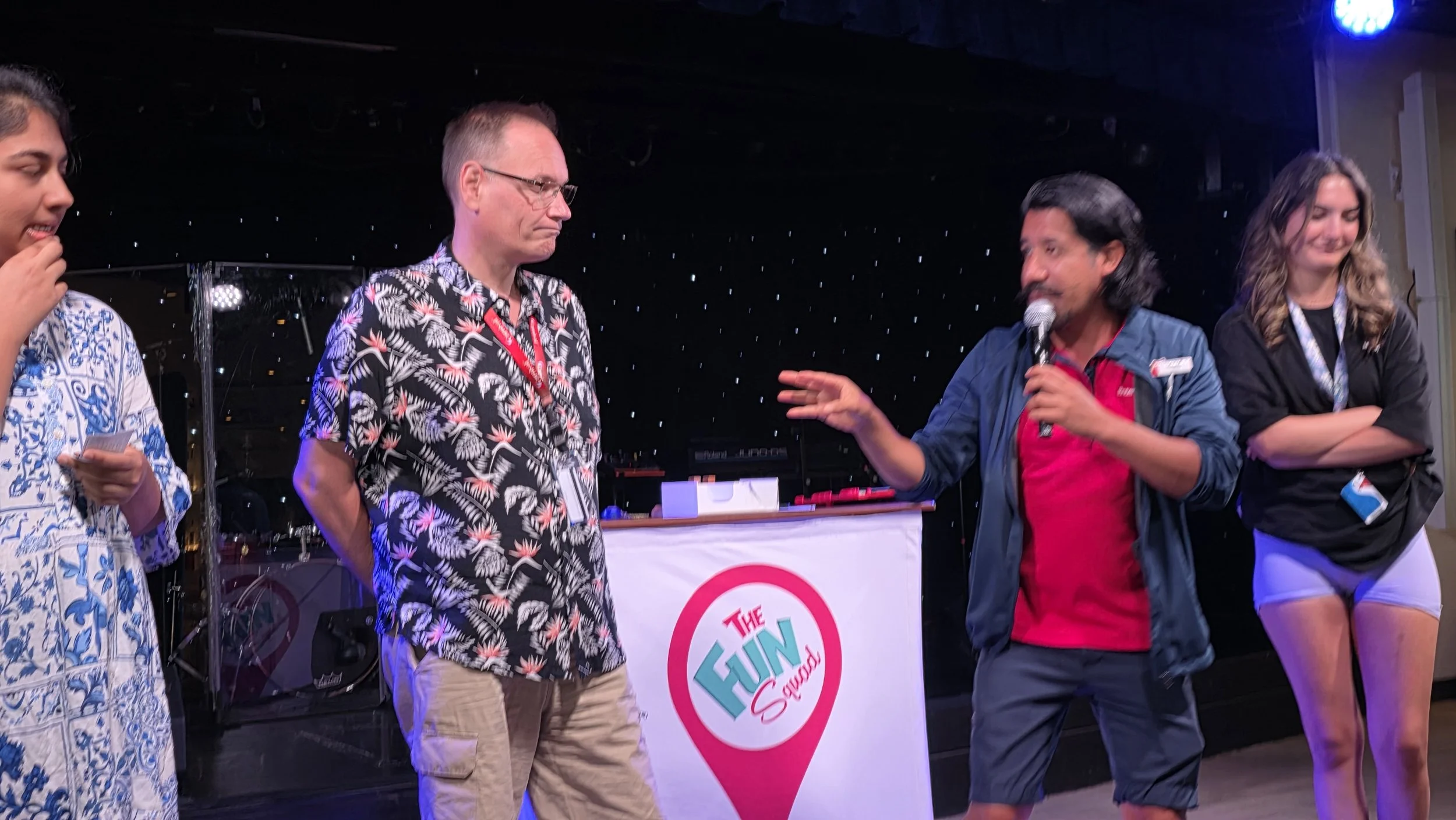 People on stage at an event called The Fun Squad, with a DJ setup in the background and a man holding a microphone talking to another man.