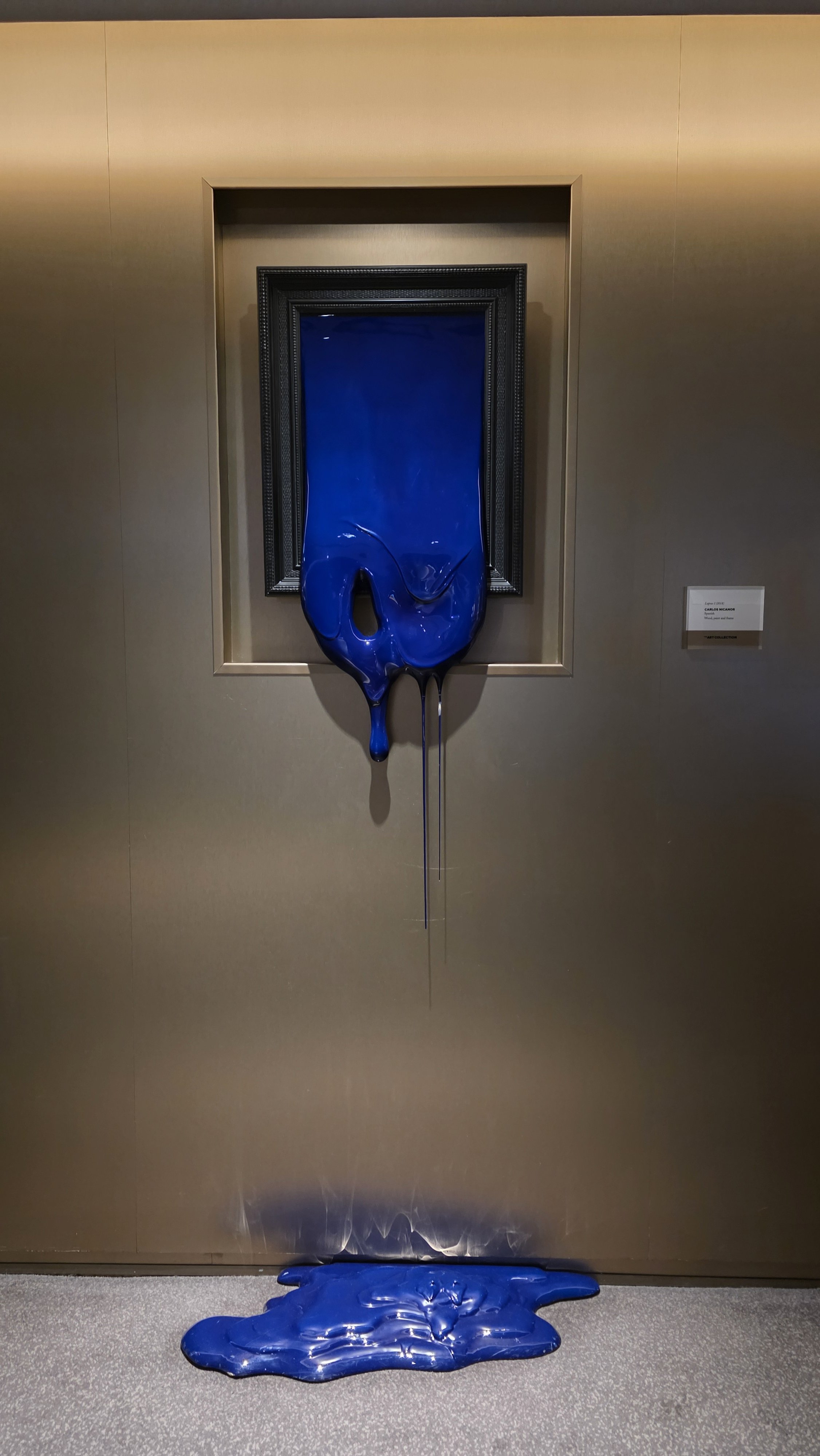 A contemporary art piece featuring a black picture frame with blue melted wax dripping down, creating a pool on the floor.
