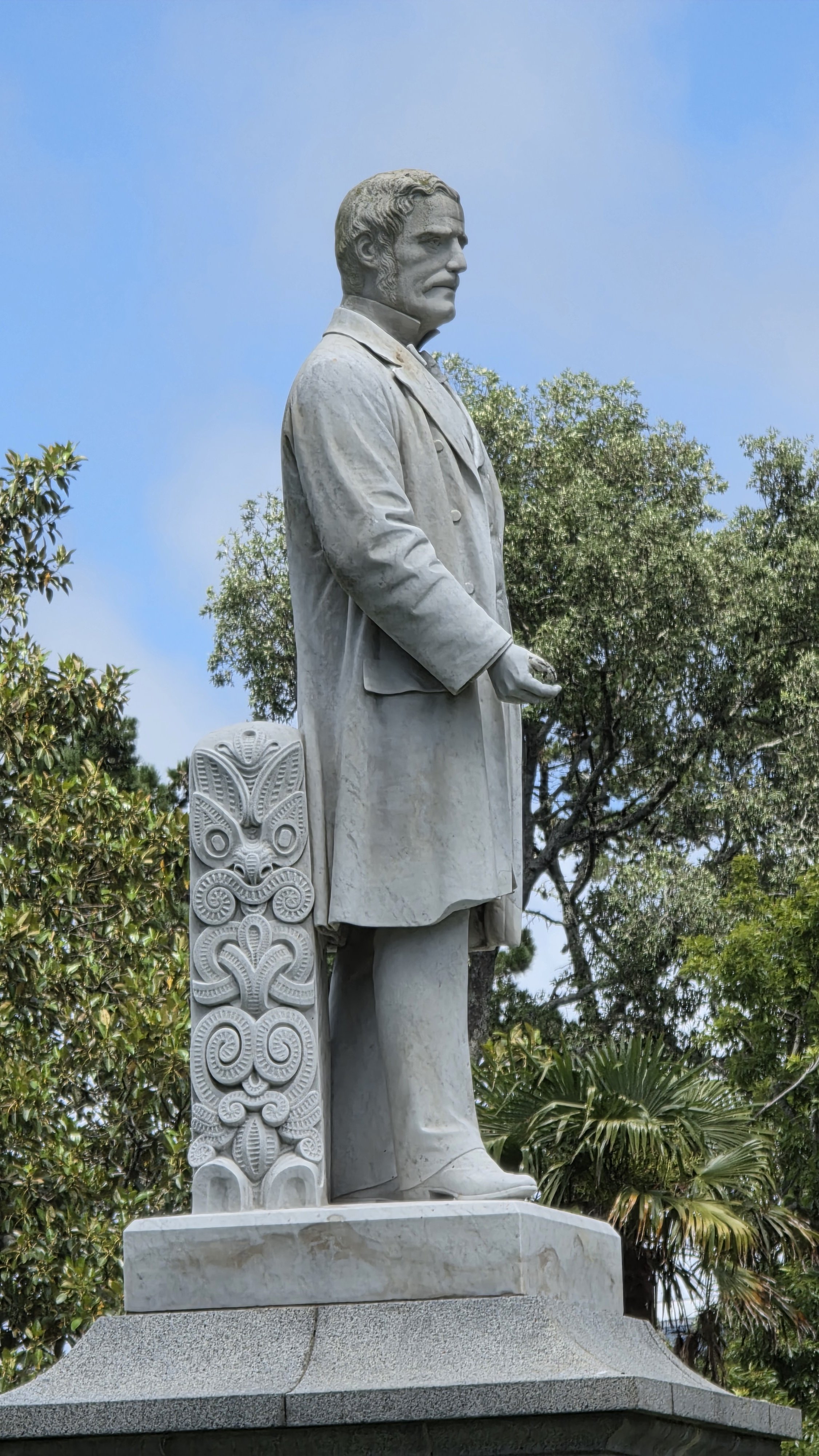 A white stone statue of a man in a trench coat and pants standing on a pedestal outdoors, with trees and a blue sky in the background.