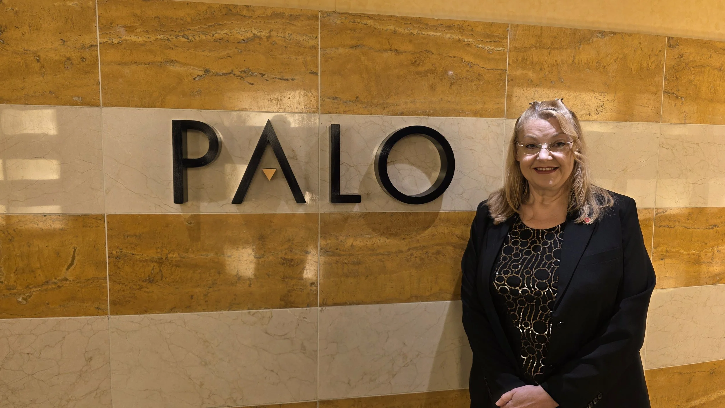 A woman with glasses and blonde hair smiling, standing in front of a wall with the word 'PALO' on it, in a building with a marble and wood interior.