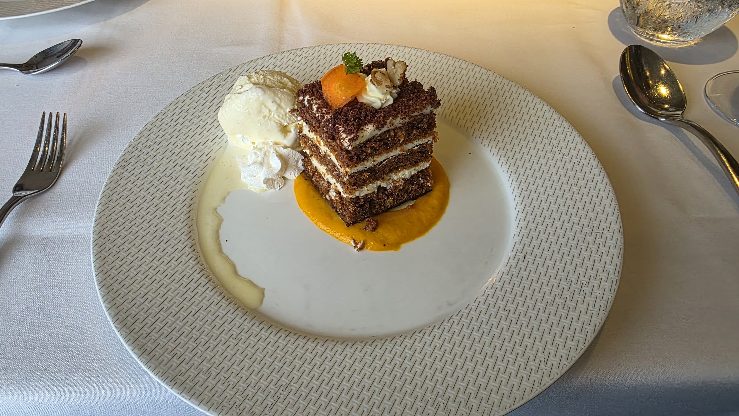 A layered chocolate cake with cream filling, topped with a small garnished orange slice and a sprig of parsley, served with ice cream and drizzle of orange sauce on a decorative white plate.