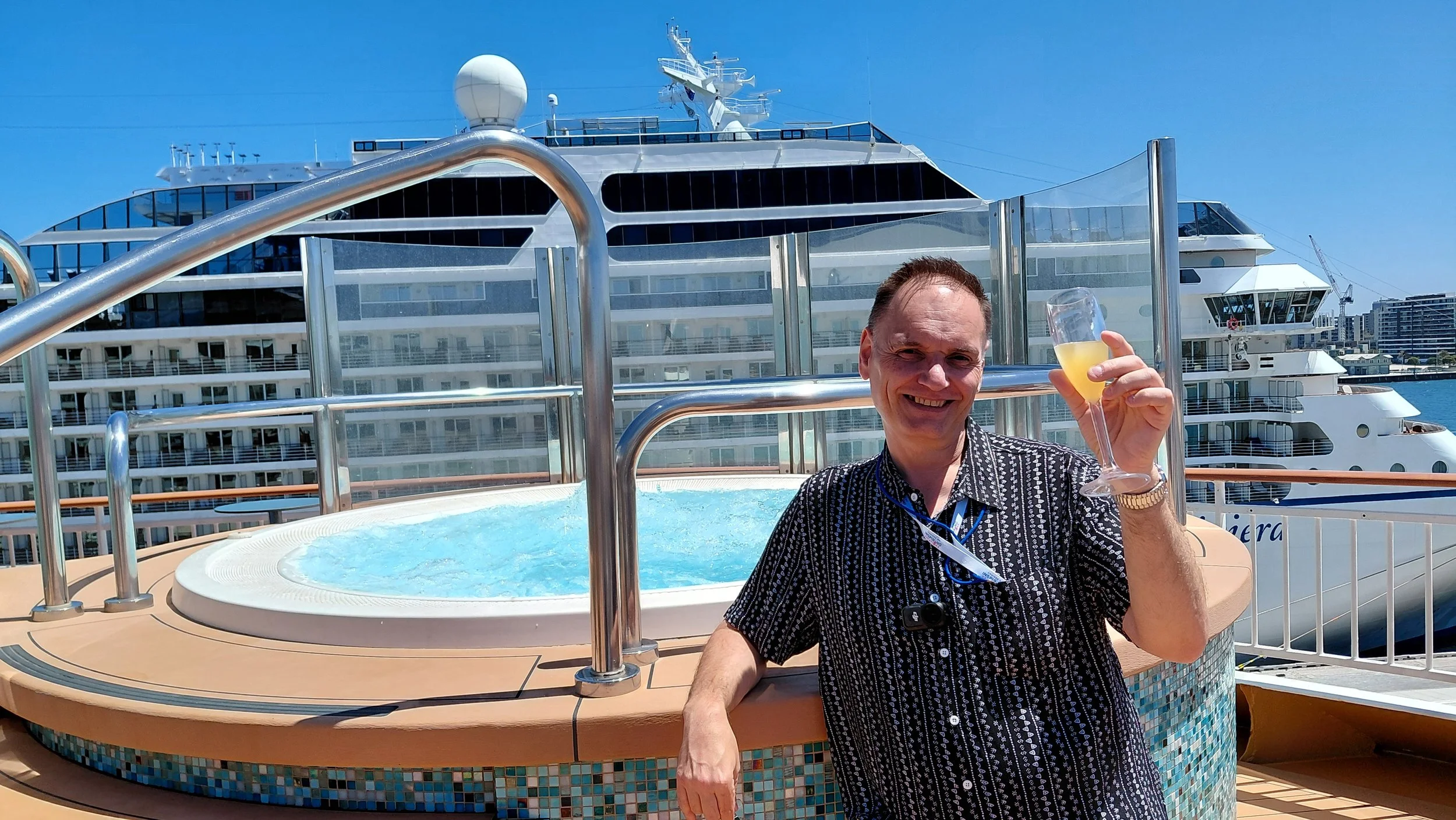 A man smiling and holding a glass of champagne on the deck of a cruise ship, with a large cruise ship in the background under a clear blue sky.