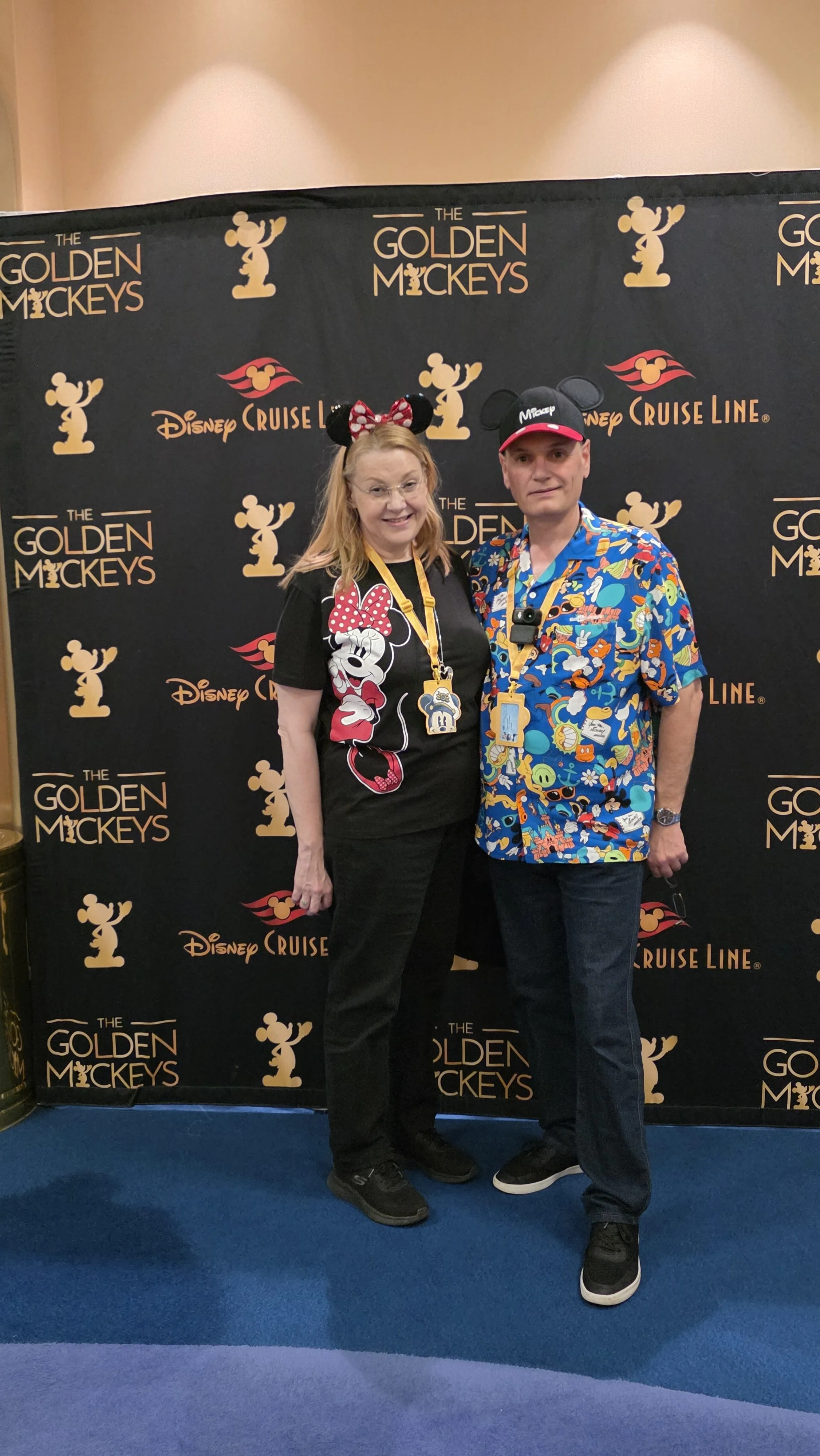 Two people standing in front of a Disney Cruise Line themed backdrop wearing Mickey Mouse ears and Disney-themed shirts.