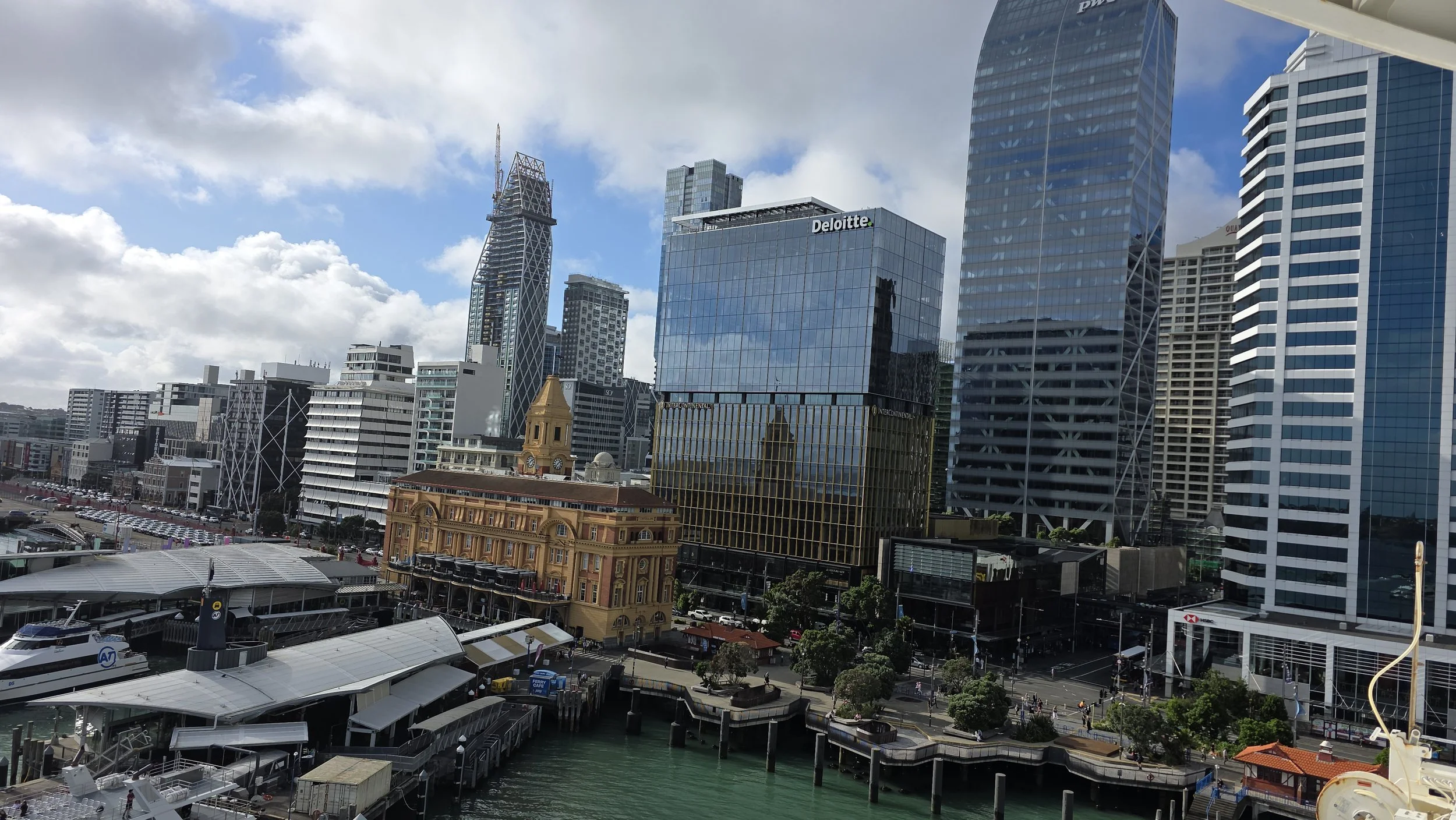 City skyline with modern glass skyscrapers, including the Deloitte building, and a historic yellow building near the waterfront with boat docks and a cloudy sky.