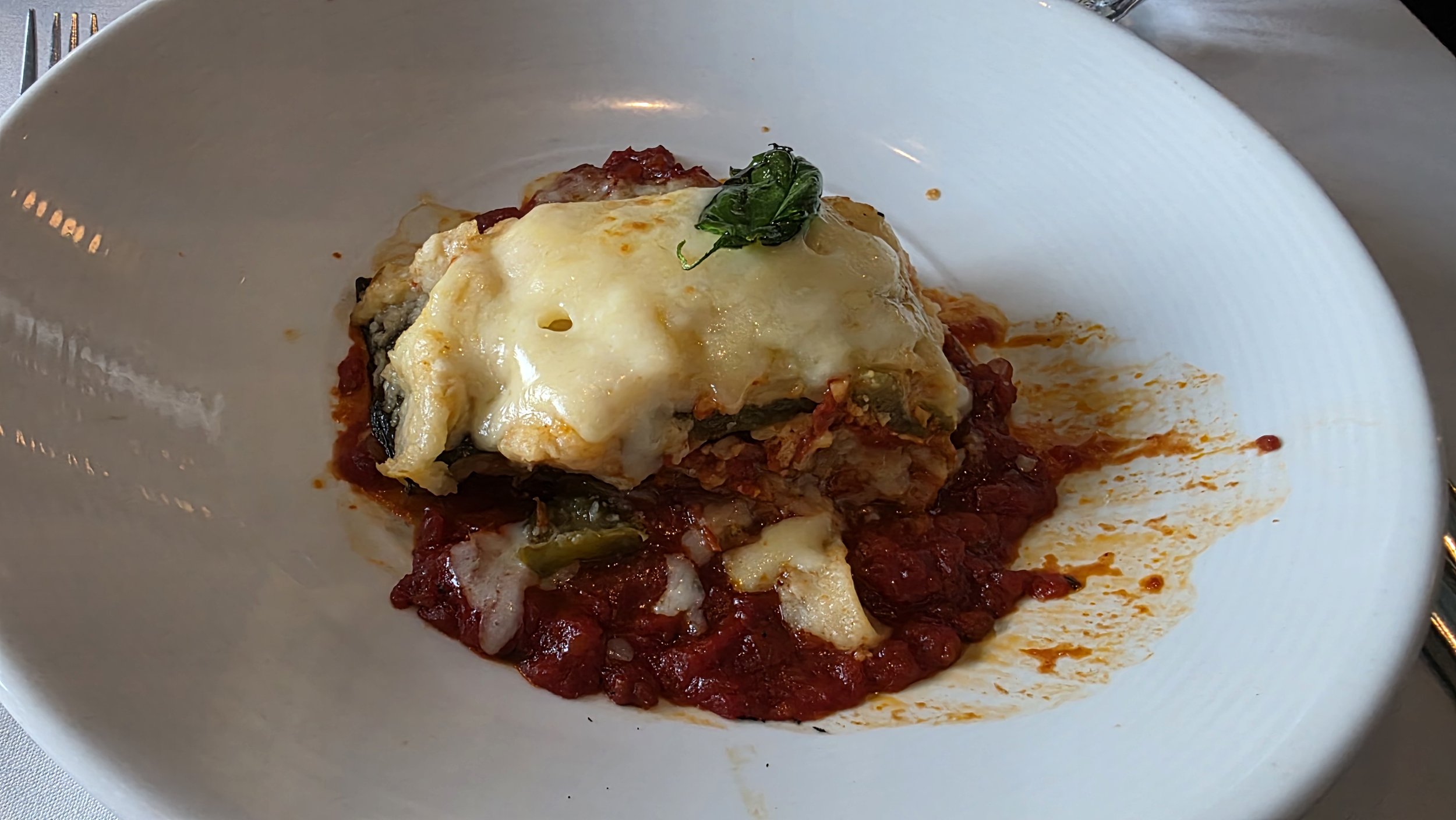 A dish with baked eggplant topped with melted cheese on a bed of tomato sauce, garnished with a basil leaf, served on a white oval plate.