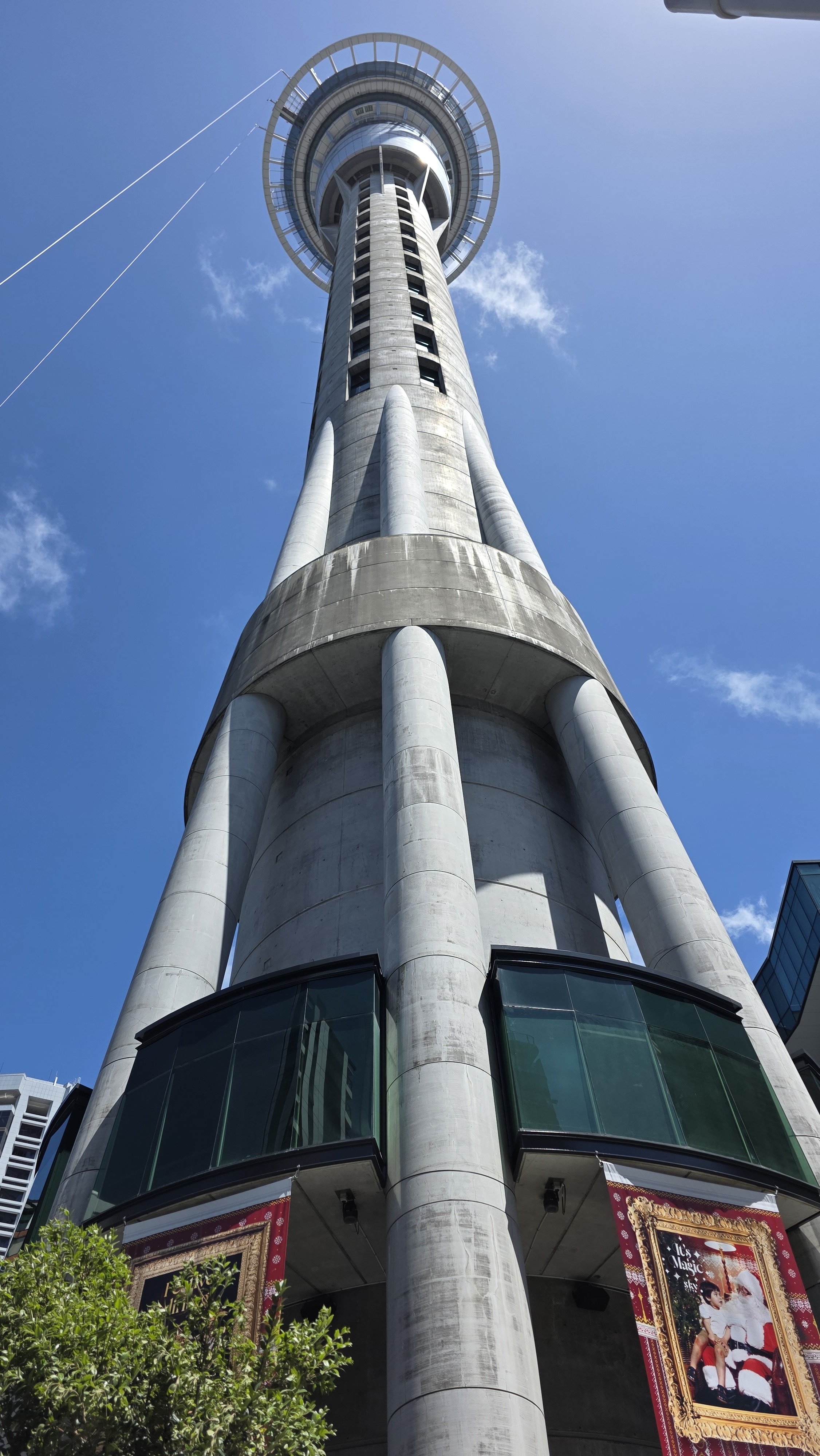 Low-angle view of Sky Tower in Auckland, New Zealand, against a blue sky with a few clouds, with a building on the right and some trees at the bottom