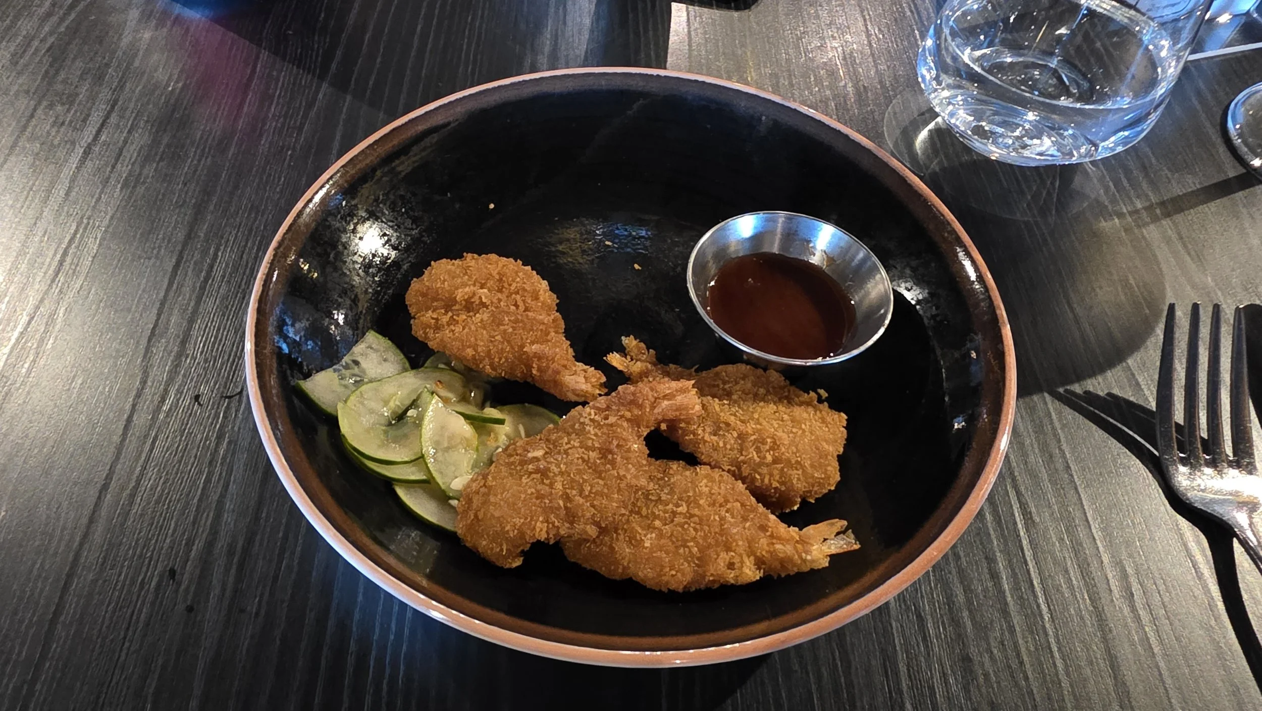 A black bowl contains fried battered shrimp, cucumber slices, and a small metal cup of dipping sauce, set on a dark wooden table.