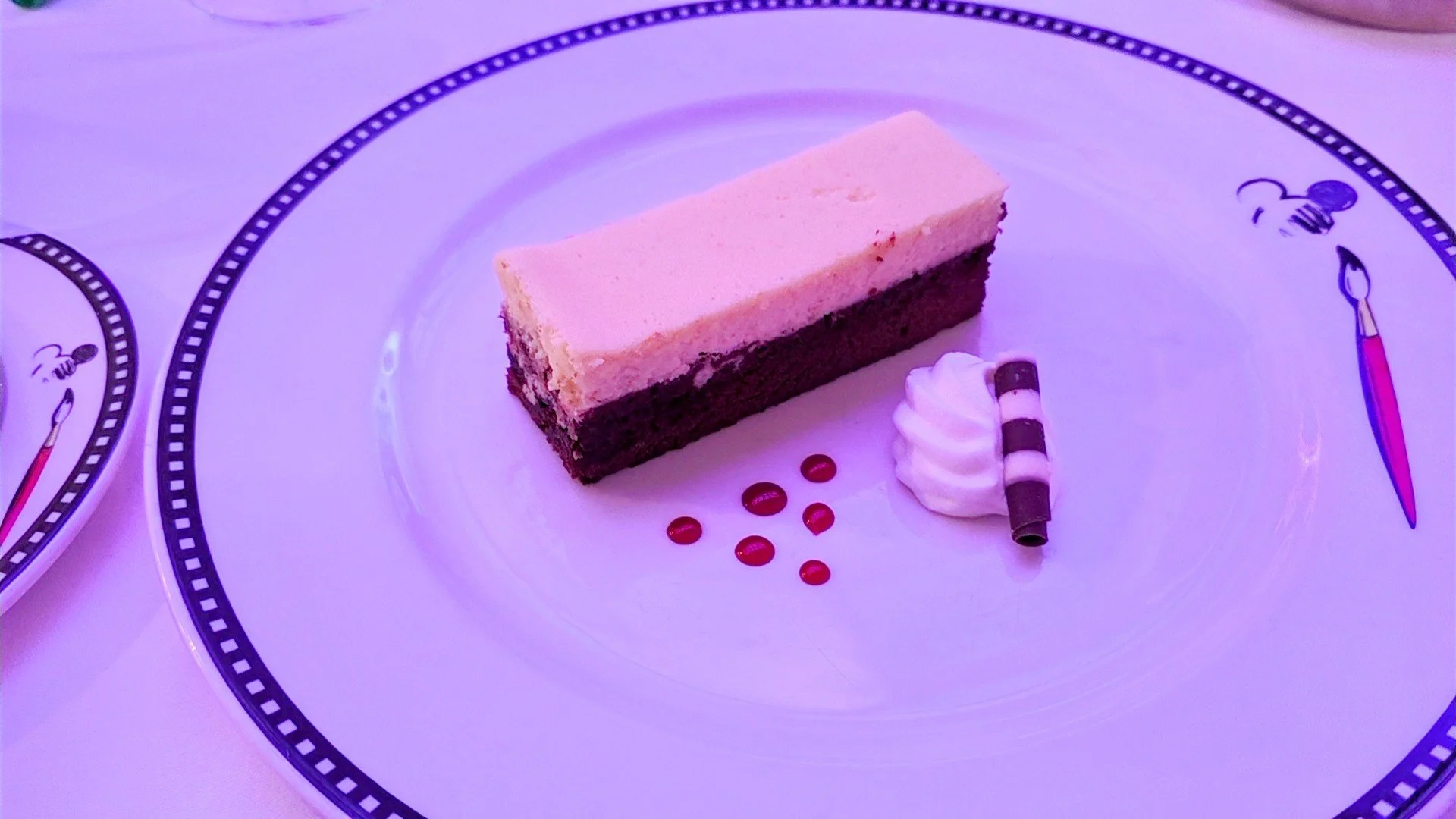 A dessert plate featuring a layered chocolate and vanilla cake slice, small red dots of sauce, a dollop of whipped cream, and a chocolate-striped wafer roll.
