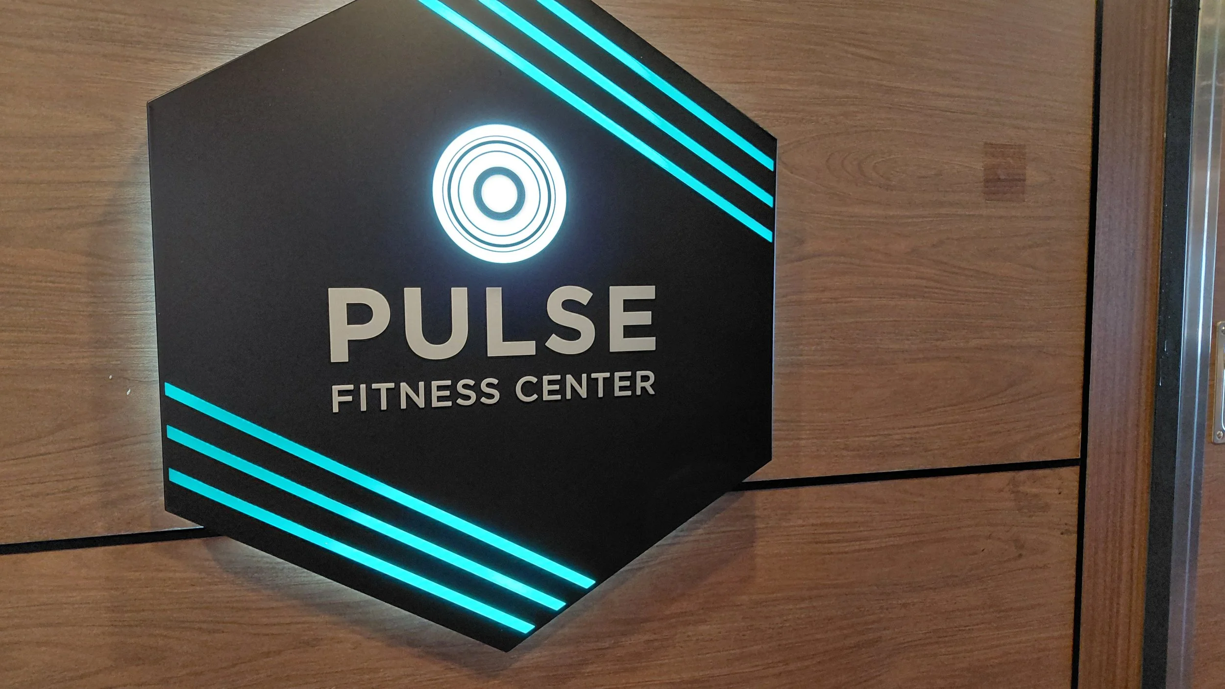 A black hexagonal sign with illuminated blue stripes and white text reading 'PULSE FITNESS CENTER' mounted on a wood-paneled wall.