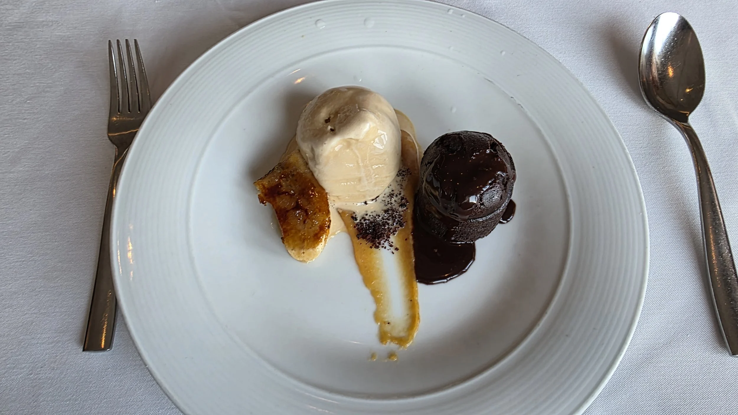 Dessert plate with a slice of caramelized banana topped with vanilla ice cream and a chocolate lava cake with melted chocolate sauce, served on a white round plate with a fork on the left and a spoon on the right.