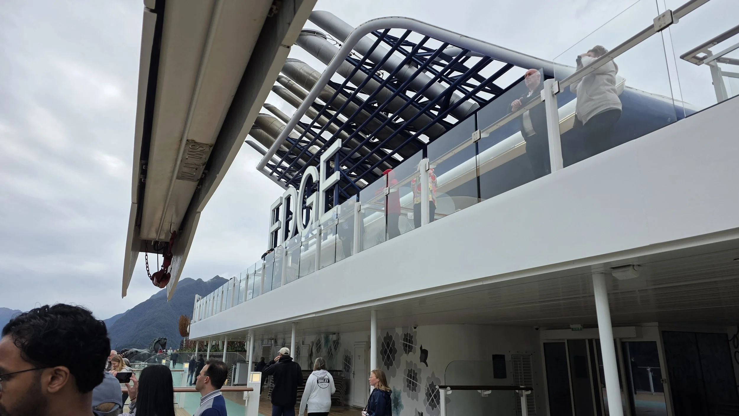 People standing on the upper deck of a ferry boat with the word 'FERRY' displayed, and mountains visible in the background.