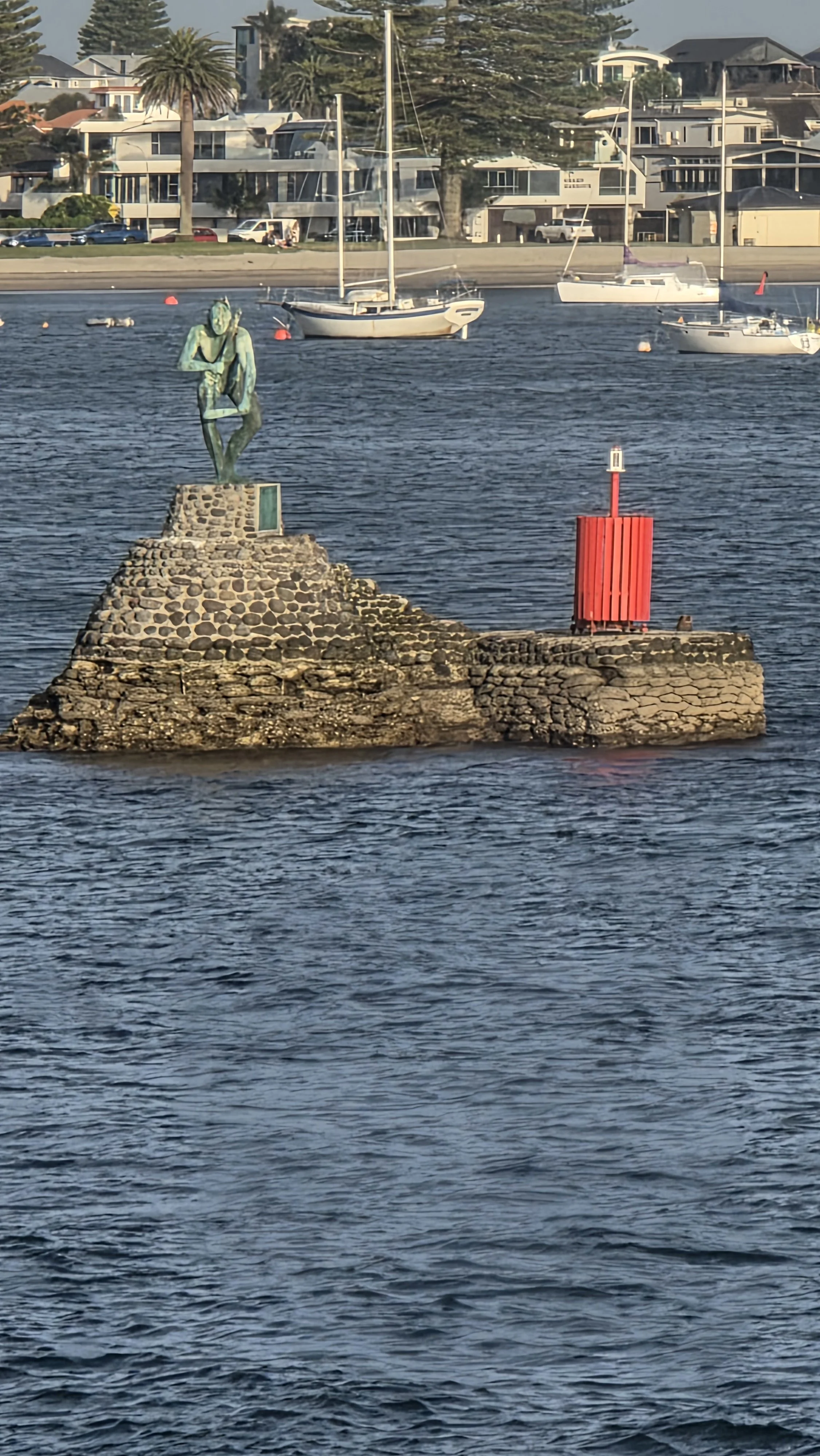 A statue of a person sitting and resting on a rock formation in the water with sailboats and residential houses in the background.