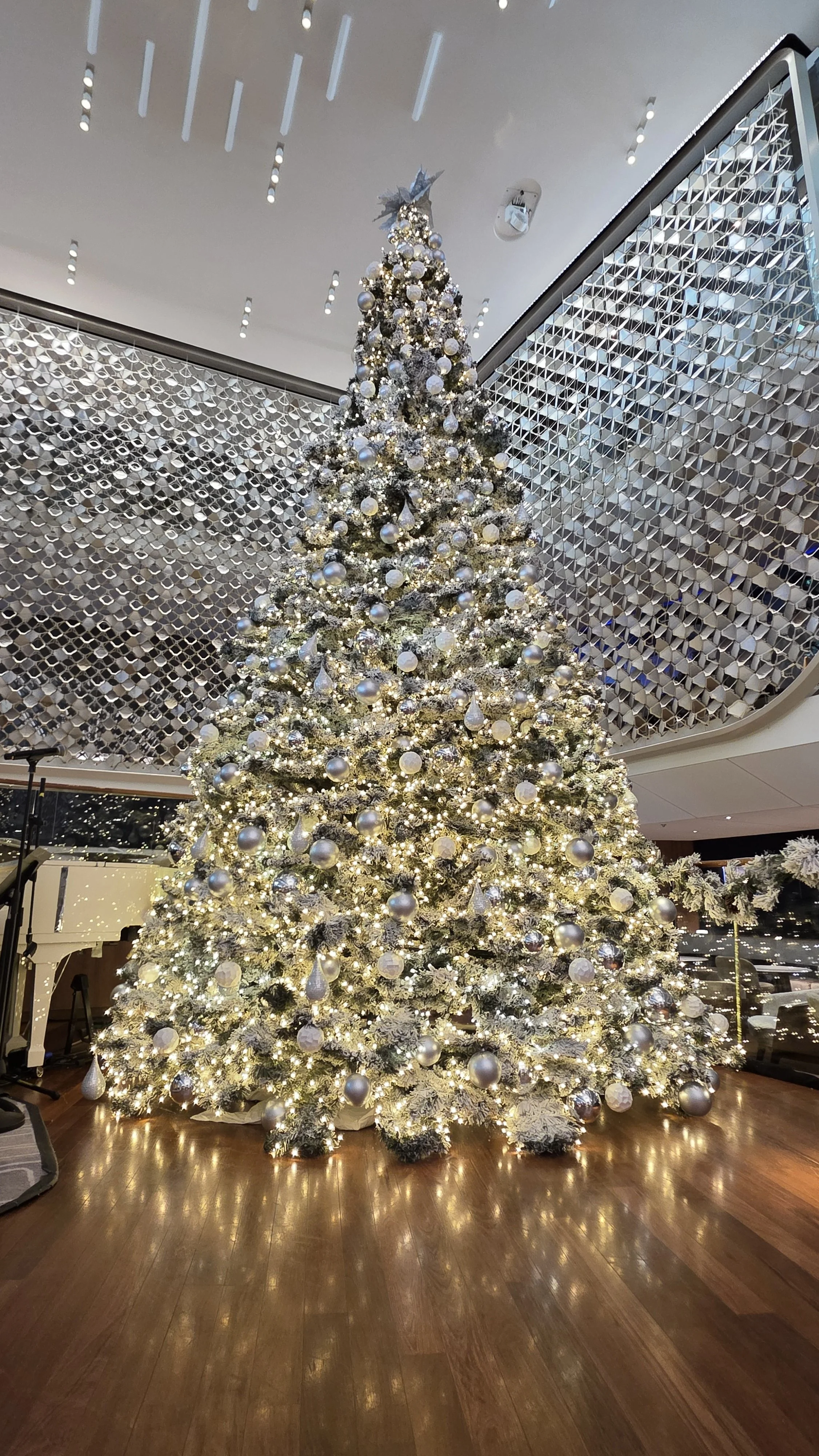 A tall Christmas tree decorated with white and silver ornaments and white lights, in a modern interior space with wood flooring and geometric wall design.