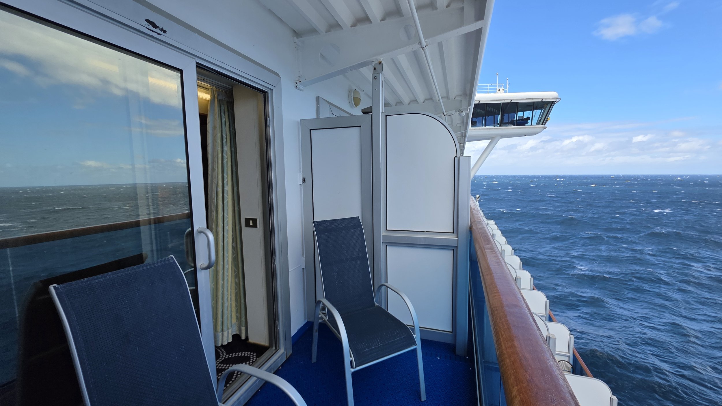 View from a cruise ship balcony showing two black chairs, a wooden railing, ocean waves, and the ship's bridge overhead in the distance.