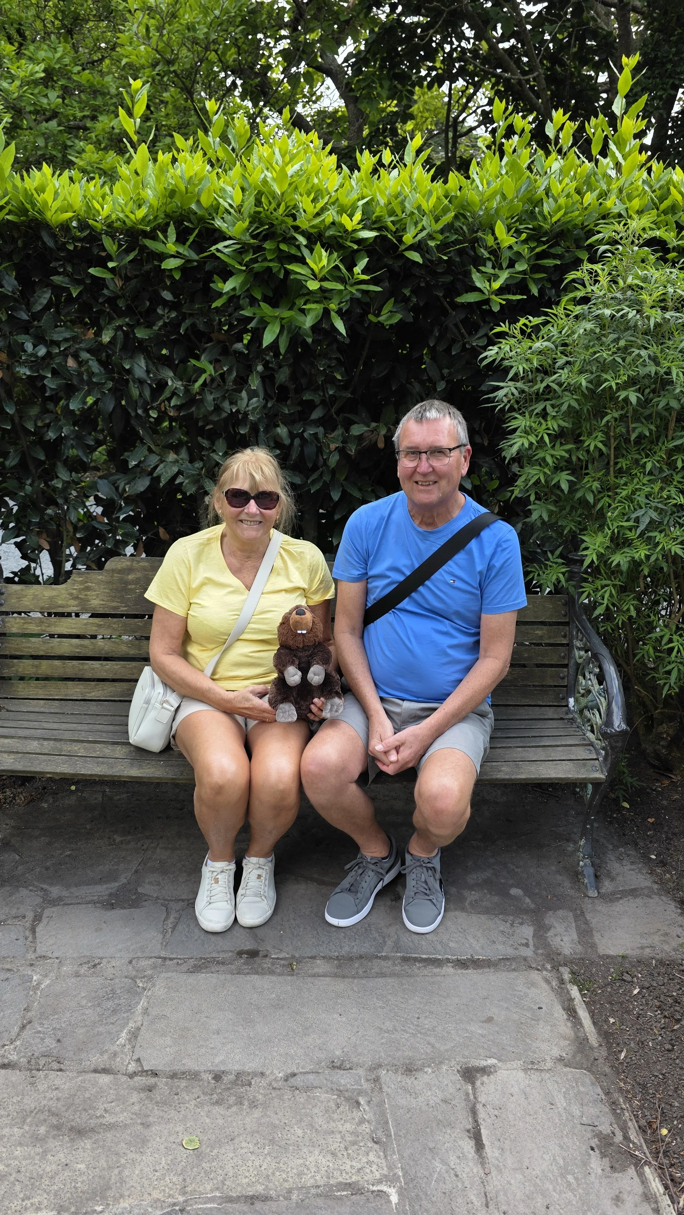 A smiling elderly woman in sunglasses, yellow shirt, and white shorts sitting on a park bench holding a plush bear, next to a smiling elderly man in glasses, blue shirt, and khaki shorts sitting on the same bench in a garden area with green foliage.