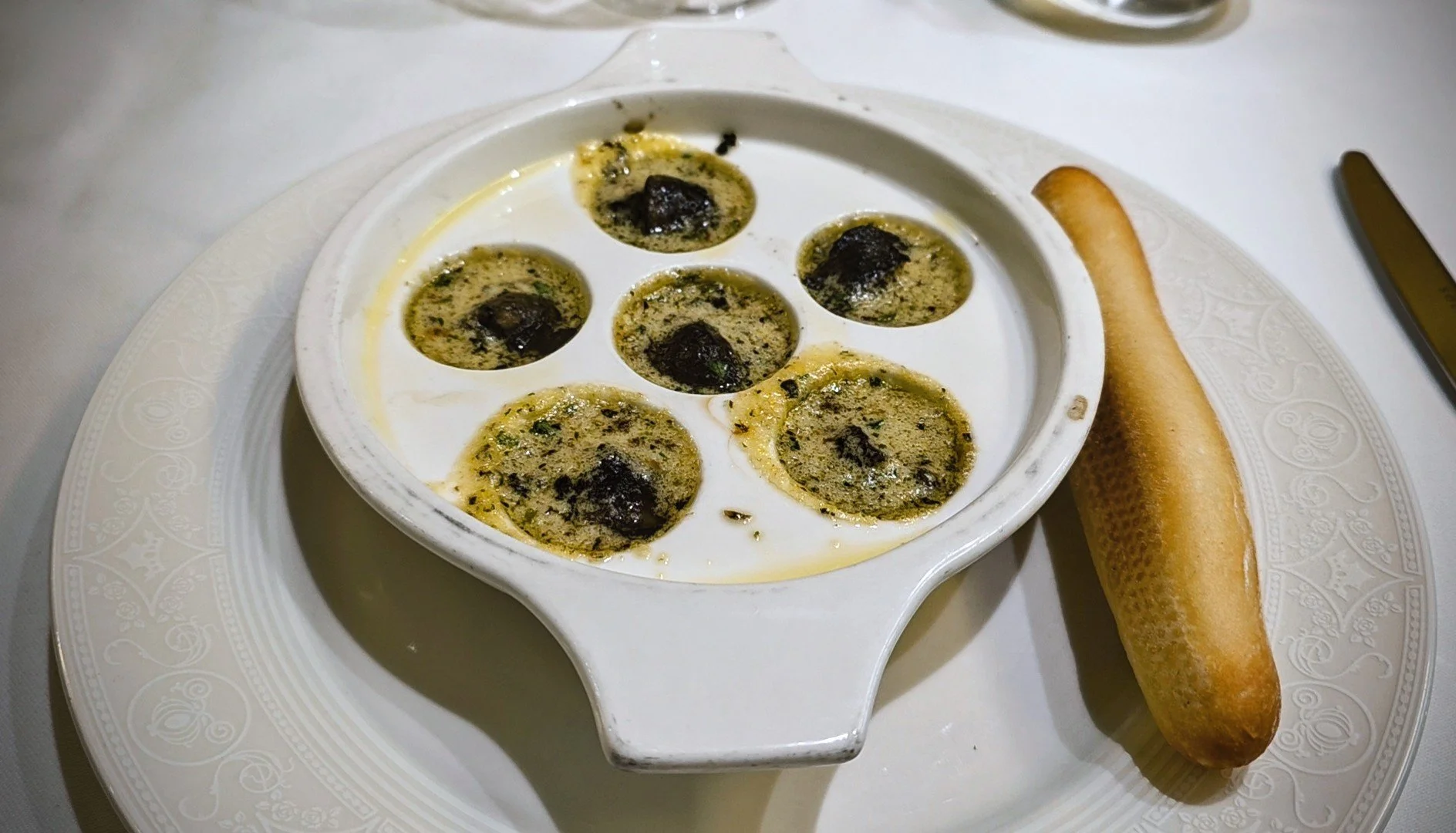 Baked escargot with herbs in a white dish, served with a breadstick on a decorative white plate.