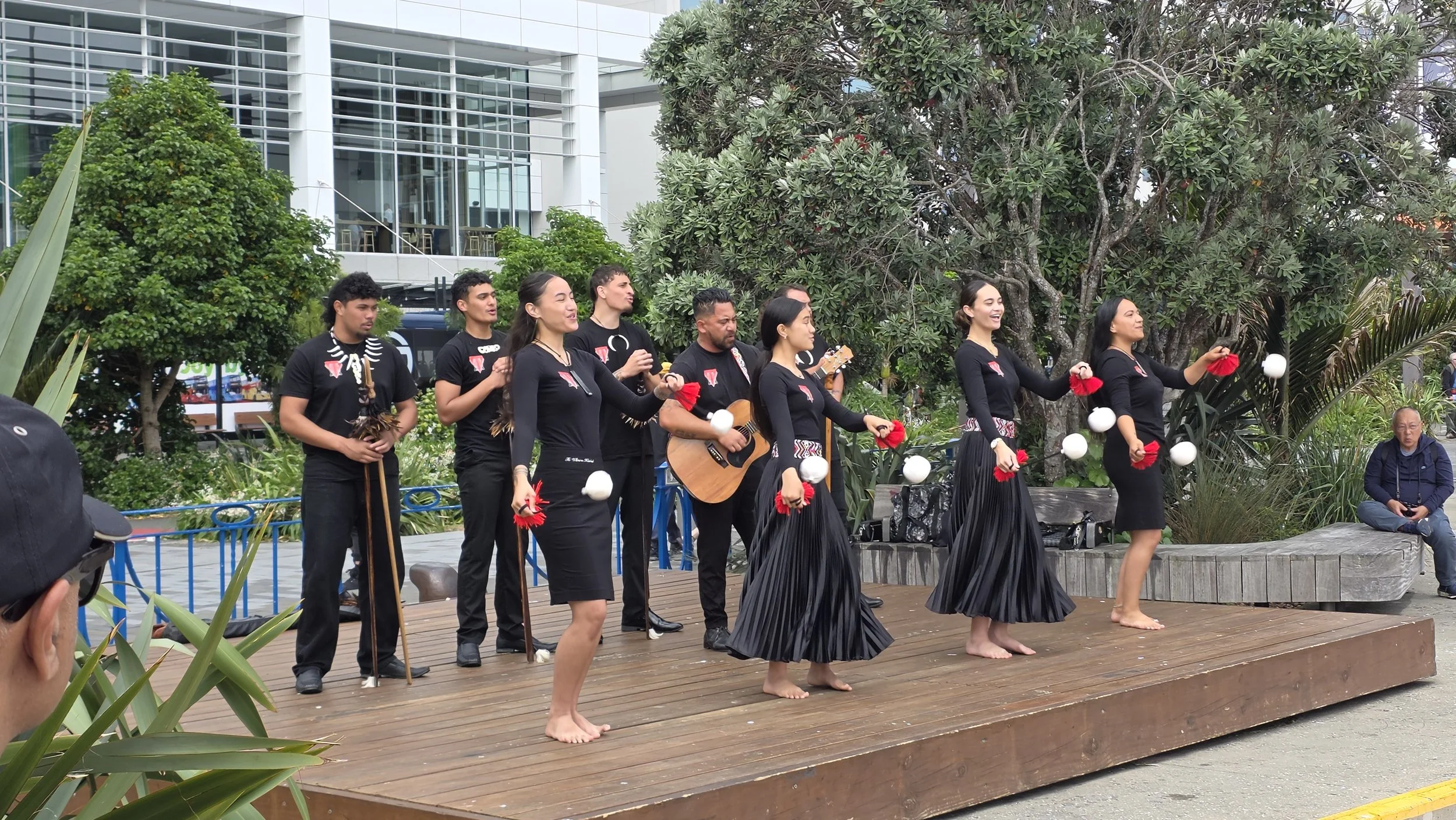 A group of performers on an outdoor stage, wearing black clothing, engaging in a cultural dance and music performance, with some playing instruments and others holding red and white props, surrounded by greenery and an audience.