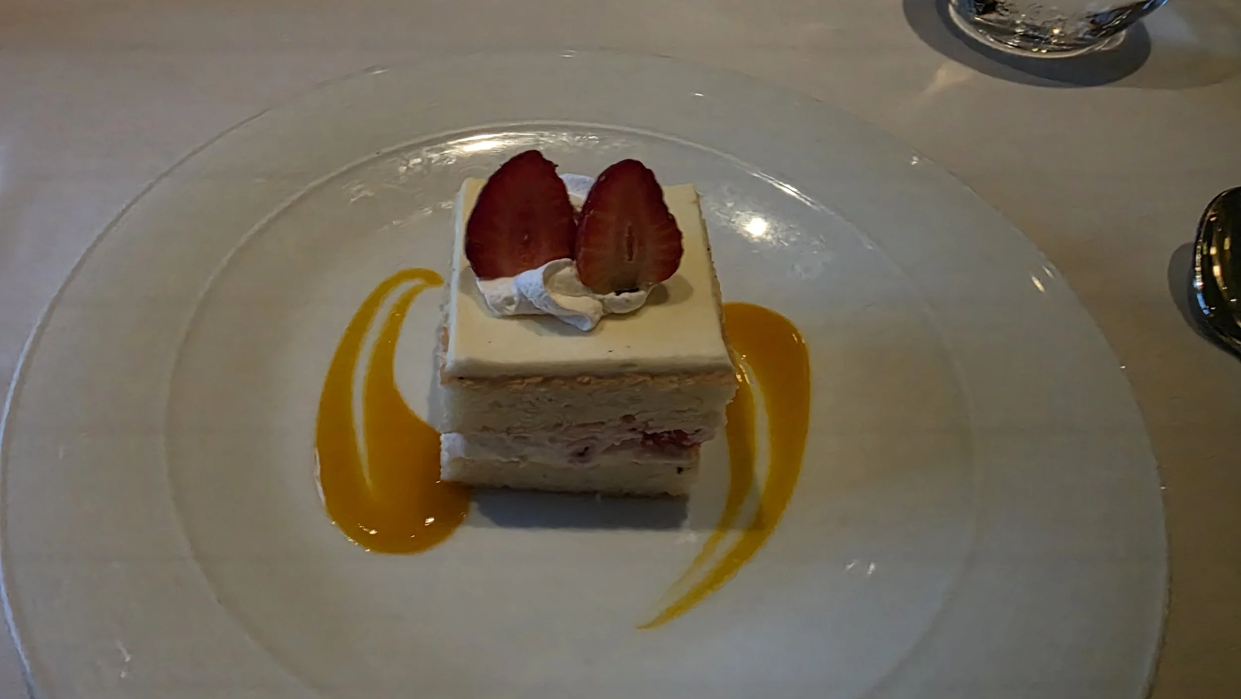 Square slice of strawberry shortcake garnished with two halved strawberries and a swirl of whipped cream, drizzled with caramel sauce on a clear glass plate.