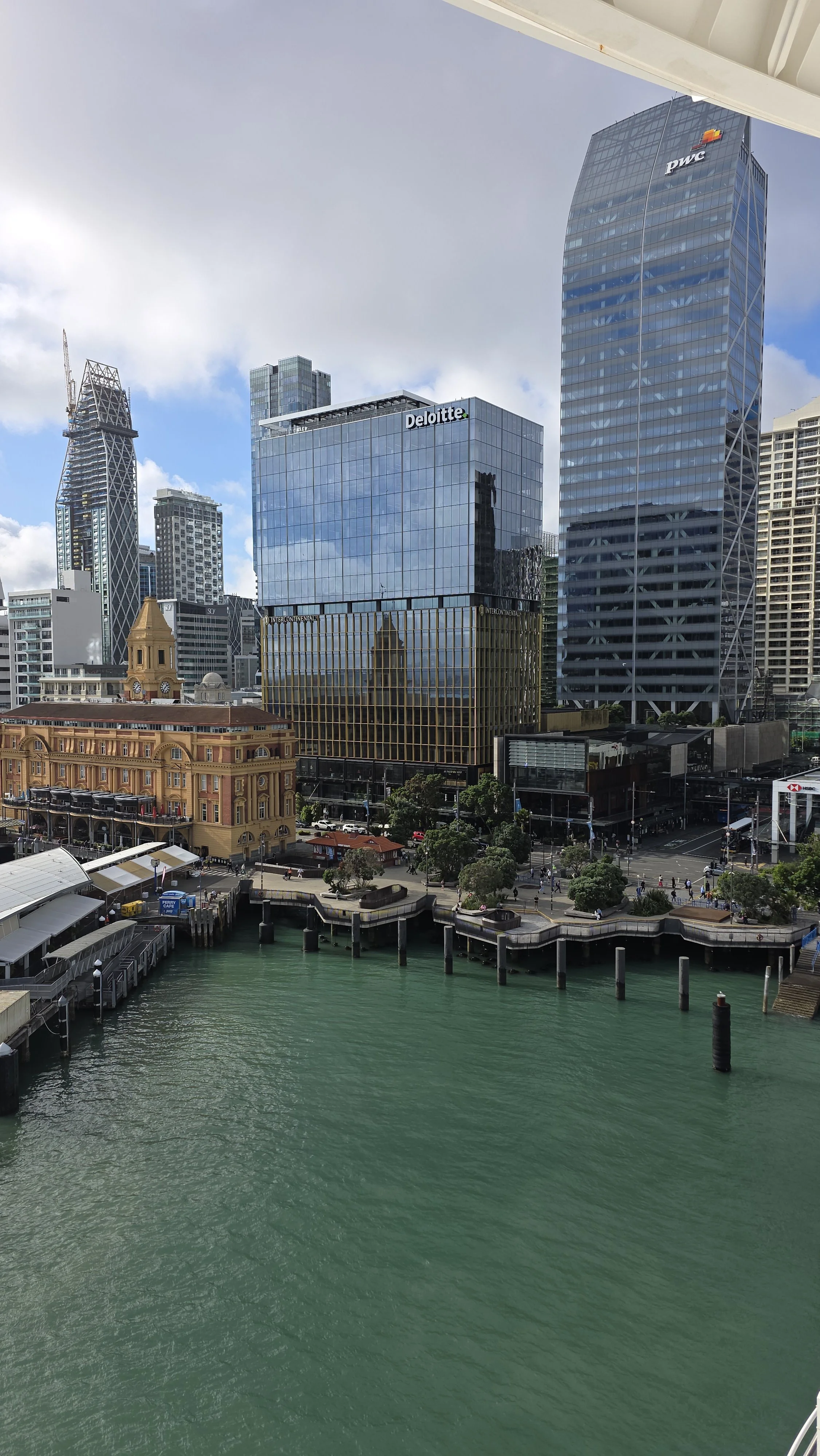 City skyline with tall glass buildings, including Deloitte and PwC, near a harbor with green water and a paved walkway along the water.