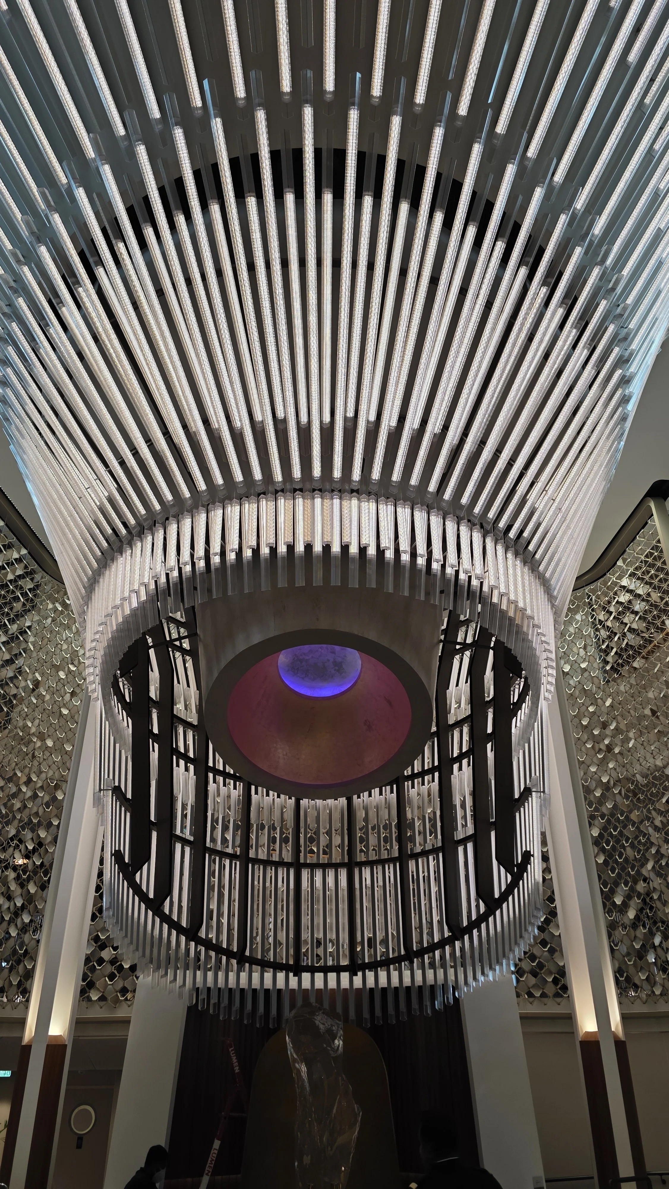Interior view of a modern building with a large, circular chandelier with vertical light tubes hanging from the ceiling, with an illuminated decorative wall and a water fountain feature below.