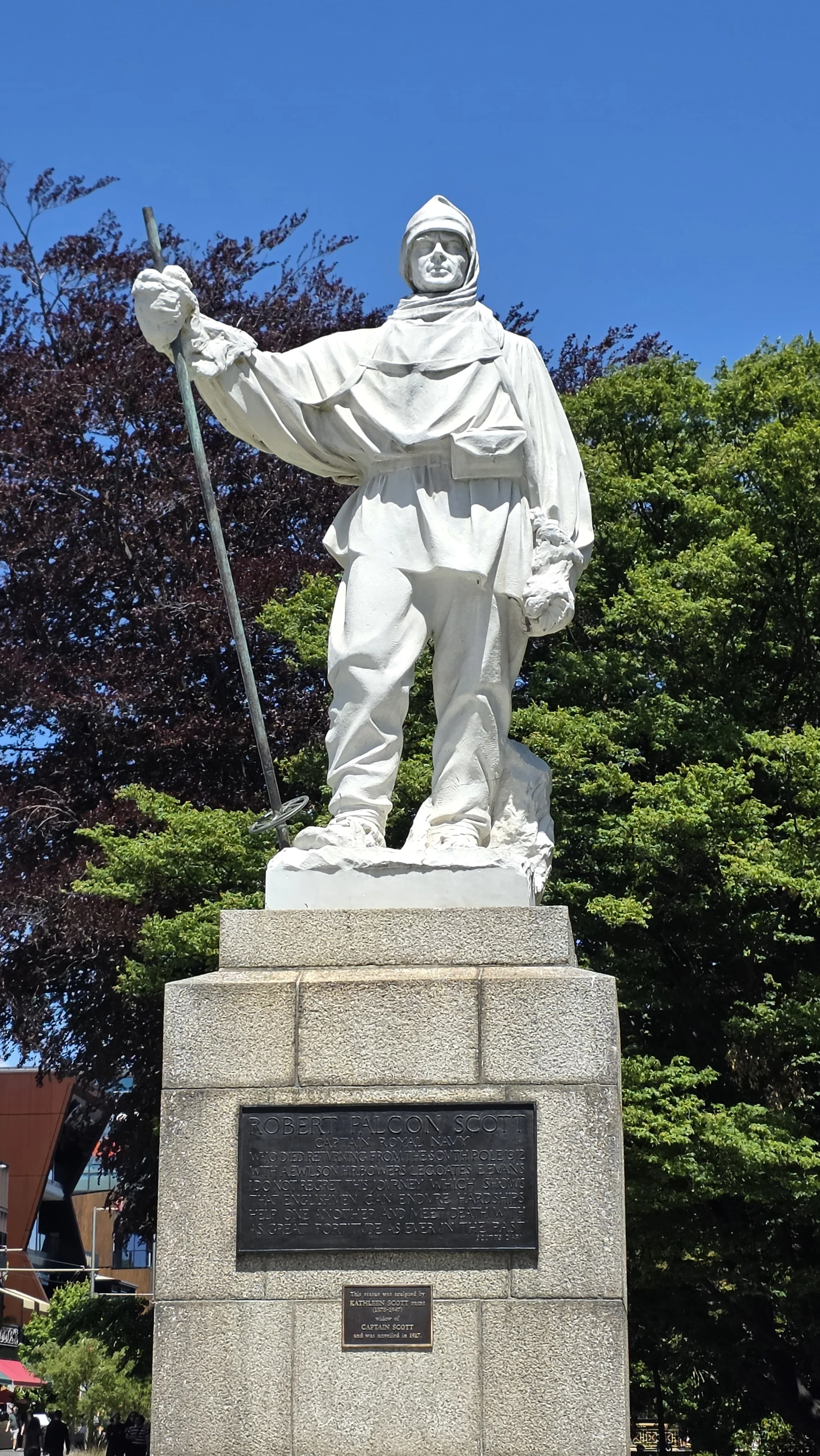 White statue of Robert Falcon Scott in military attire holding a rifle with the right hand, standing on a stone pedestal in a park with trees in the background.