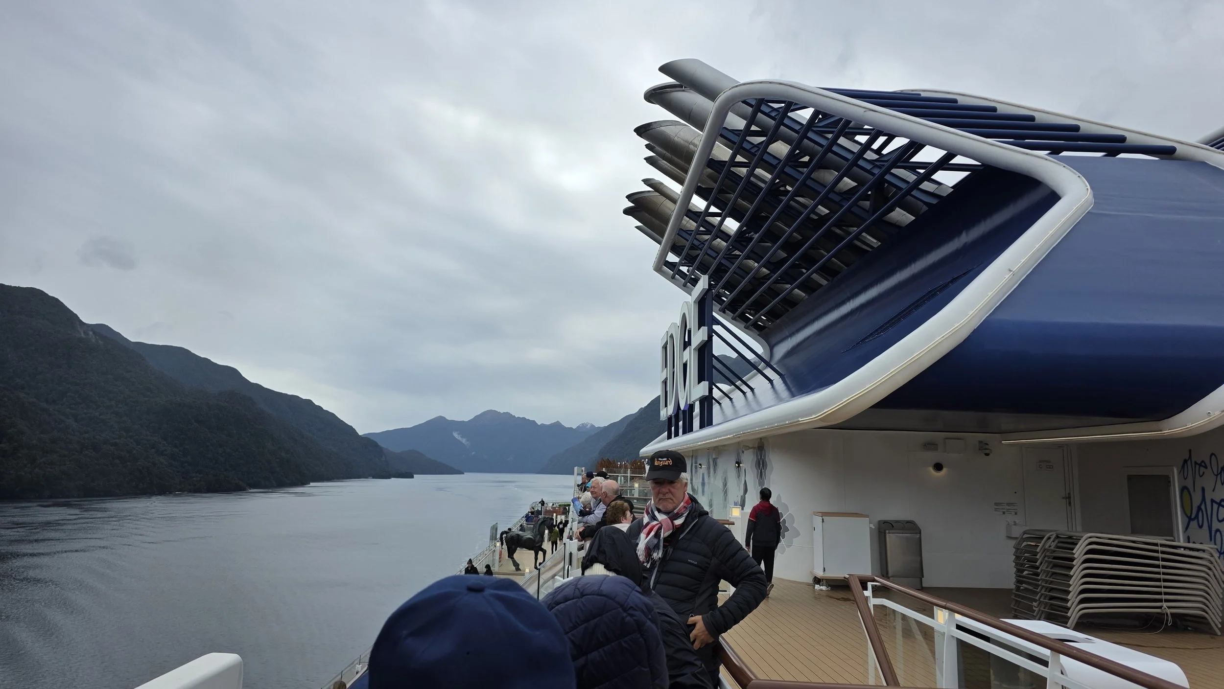 People standing and walking on the deck of a cruise ship, with mountains and a calm waterway in the background.
