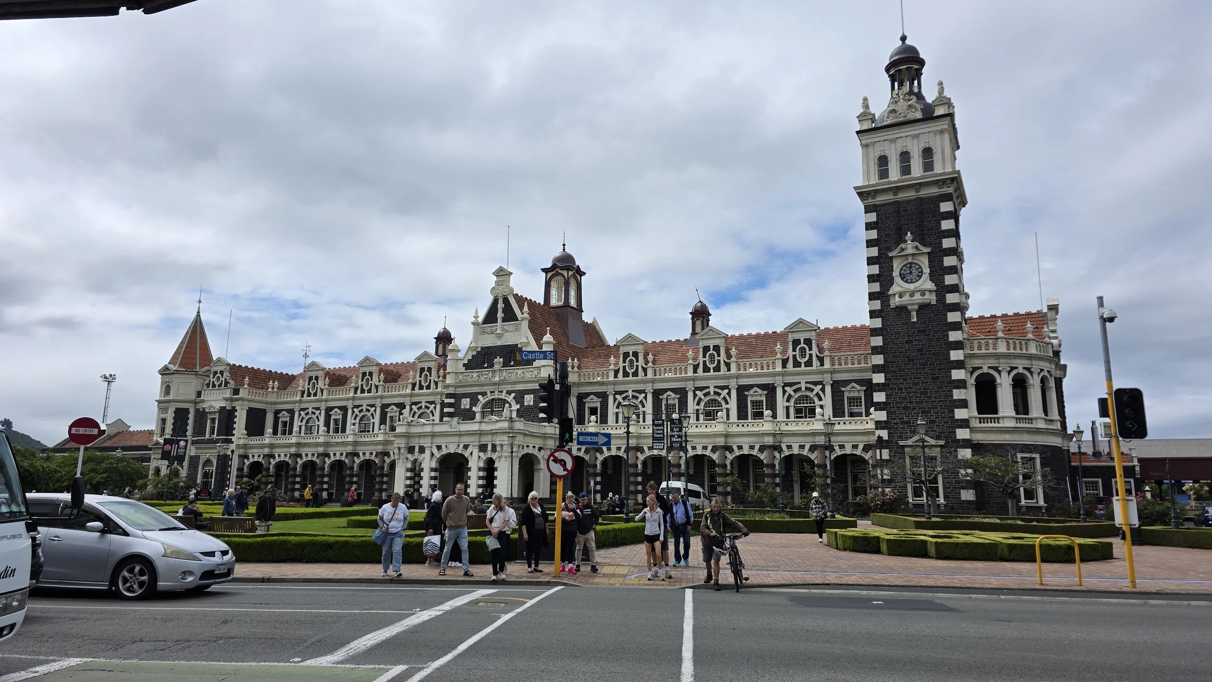 Historic building with clock tower and people walking on the sidewalk in front, cars parked nearby, cloudy sky.