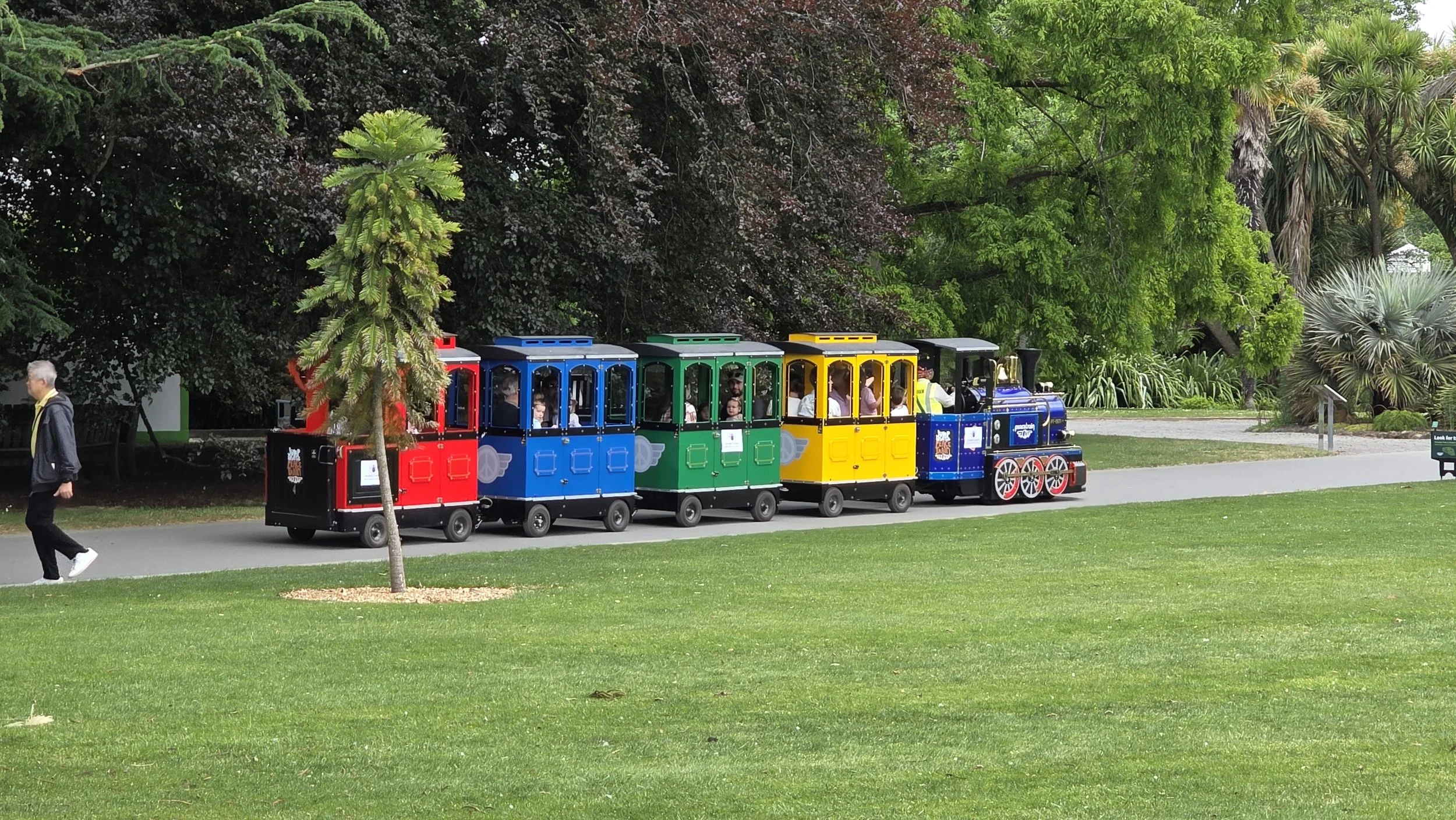 Colorful miniature train with red, blue, green, yellow, and blue engine cars moving through a park with green trees and grass.