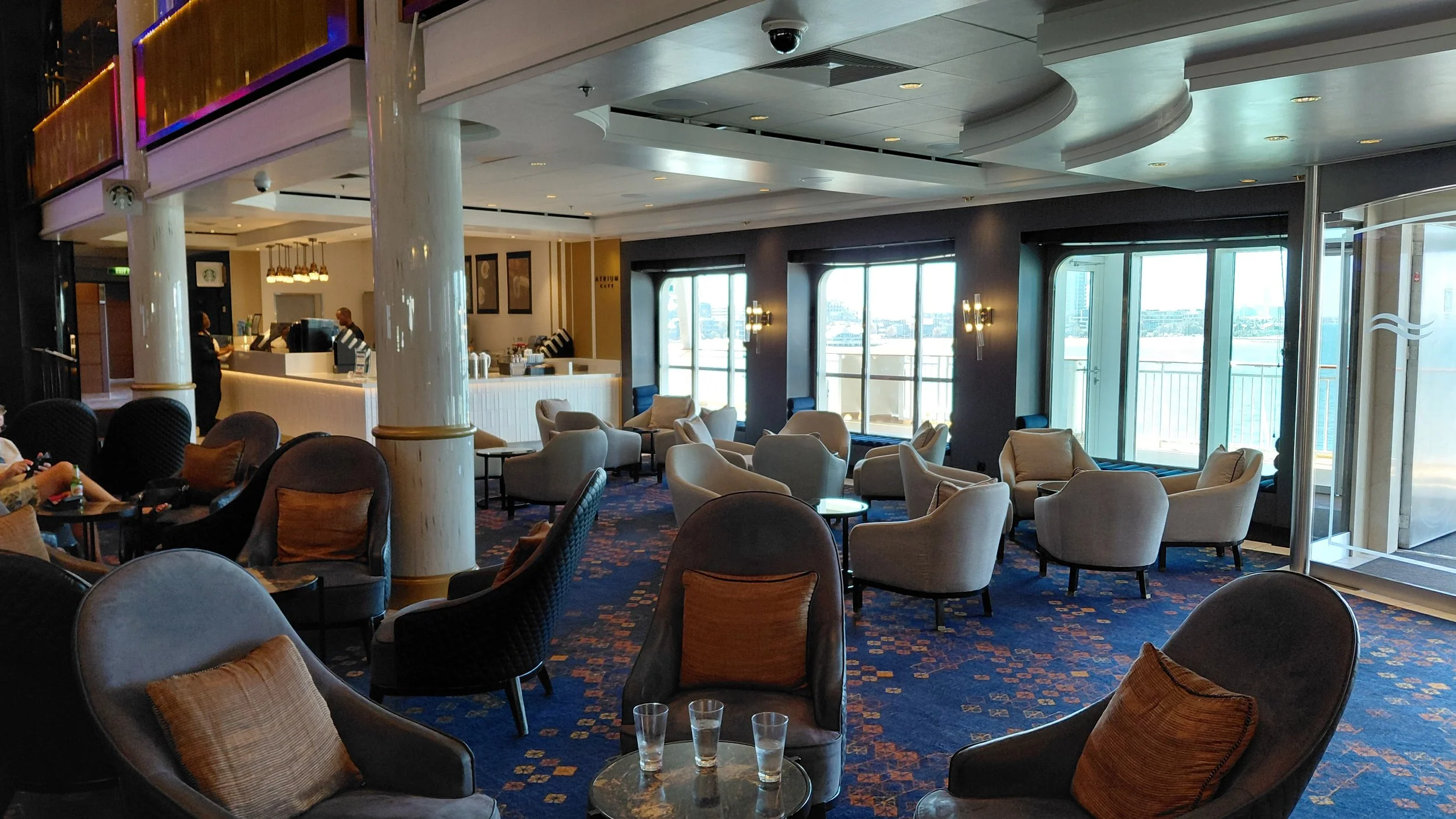 An interior view of a lounge area with several armchairs, small tables, and large windows with views outside. There are people at a bar in the background and the space has a modern, elegant design with dark walls, gold accents, and patterned carpet.