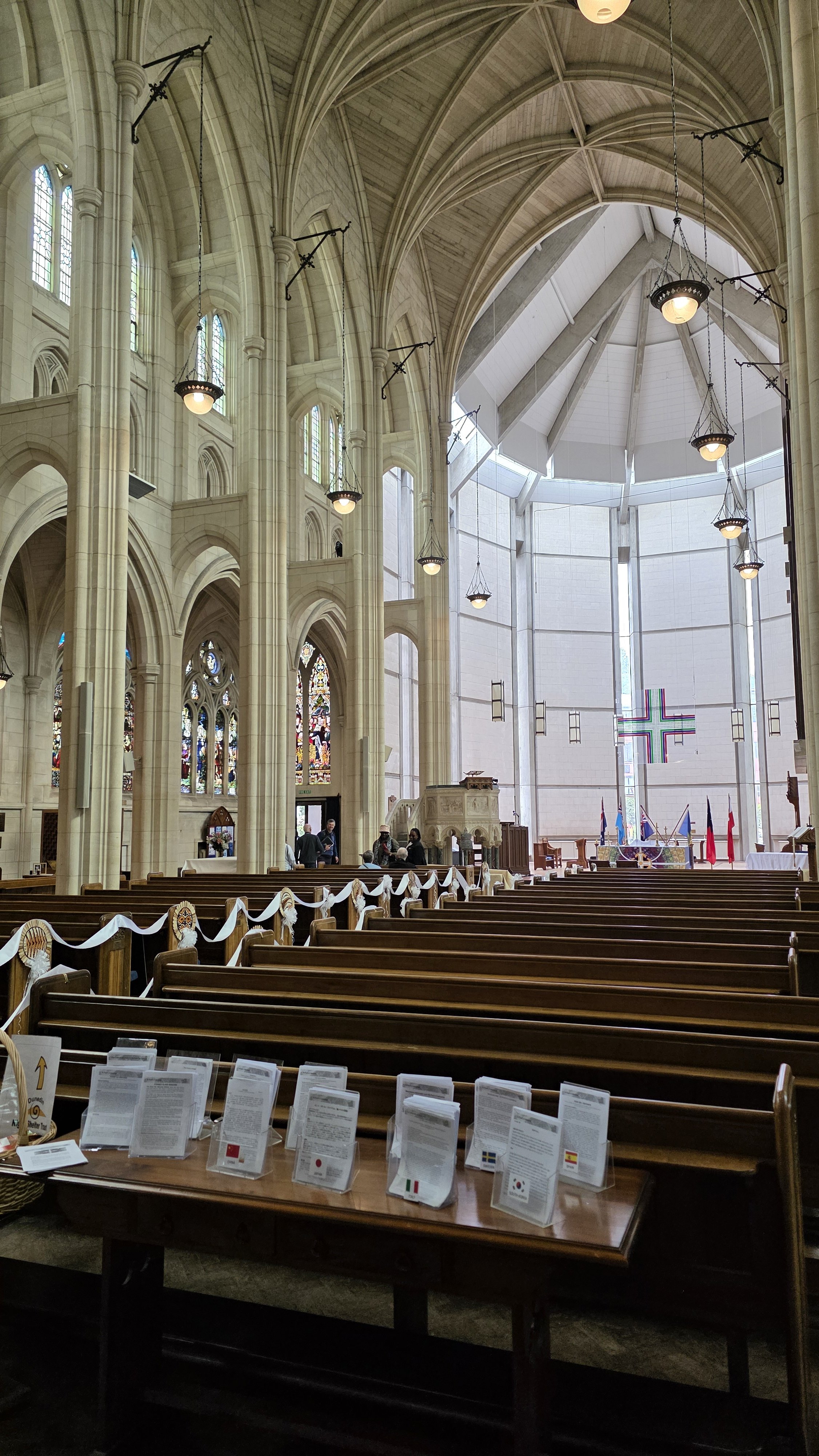 Inside a large Gothic-style church with high vaulted ceilings, stained glass windows, and wooden pews decorated with white ribbons. The altar area has a cross, flags, and a few people present.