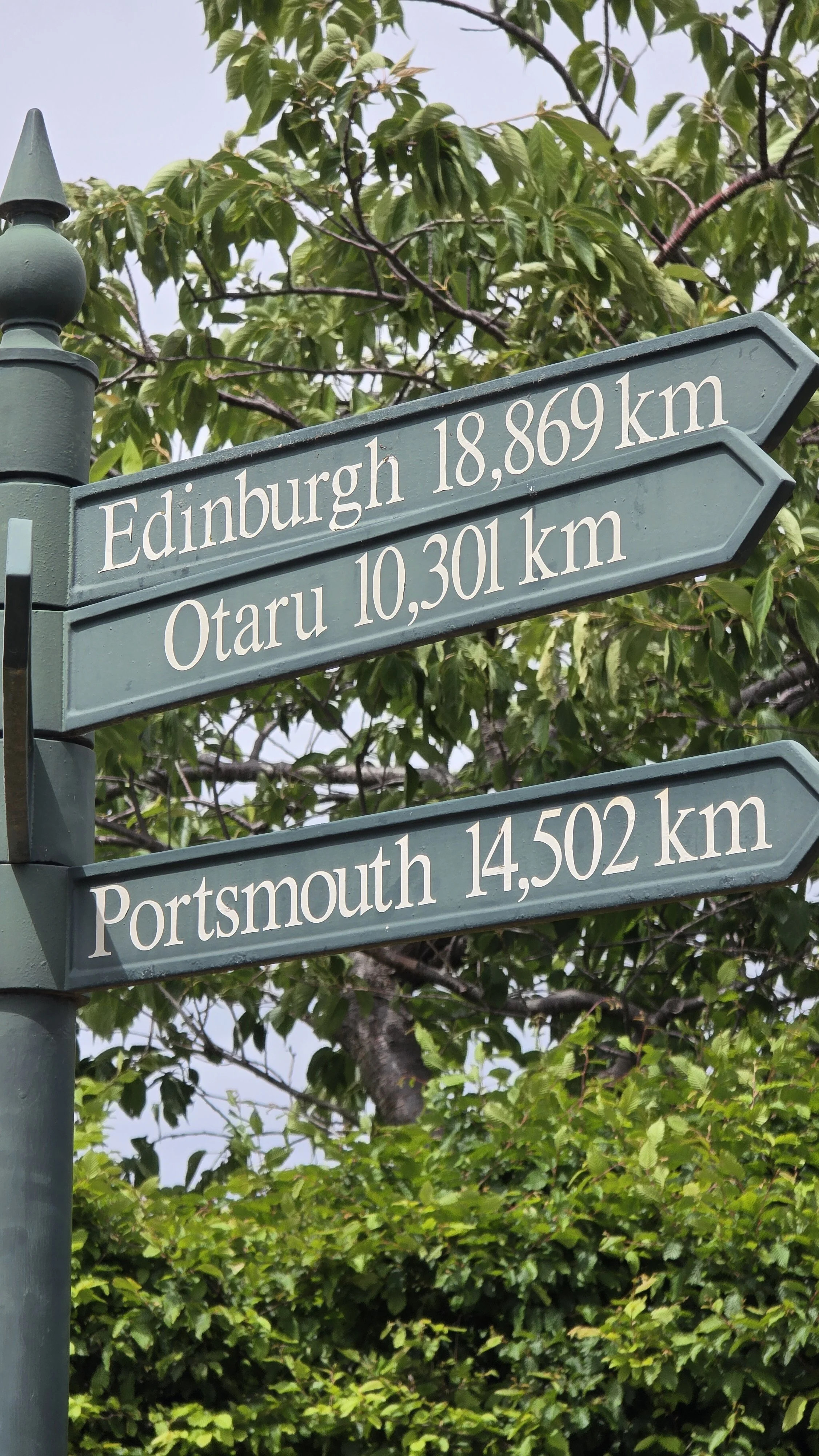 Street sign showing distances to Edinburgh, Otaru, and Portsmouth, with trees and sky in the background.