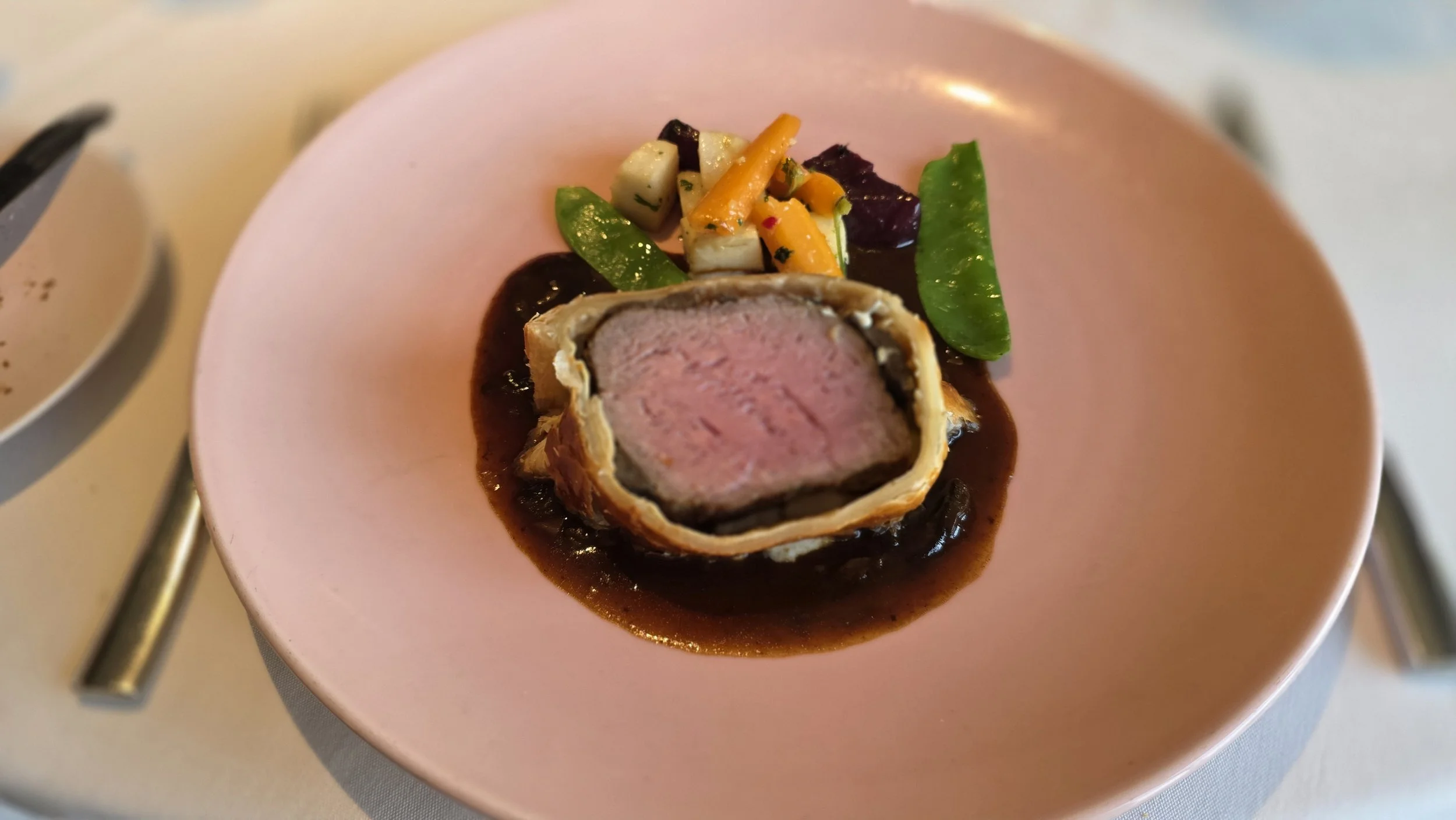 A plated gourmet dish with a portion of beef Wellington on a pink plate, accompanied by vegetables and a dark sauce.