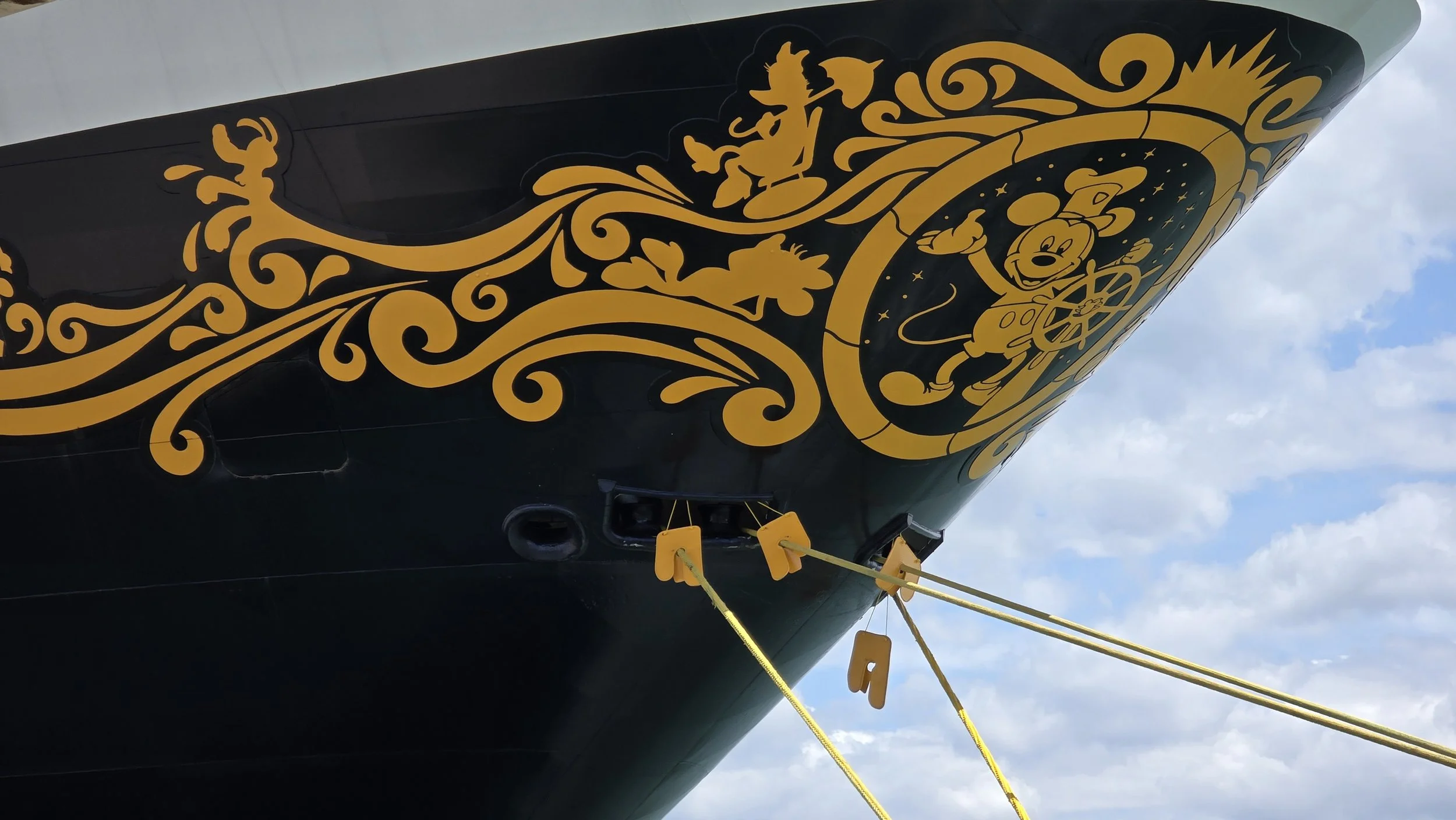 Close-up of the bow of a Disney cruise ship with black and gold decorative art featuring Mickey Mouse and other Disney characters, with yellow mooring ropes attached.