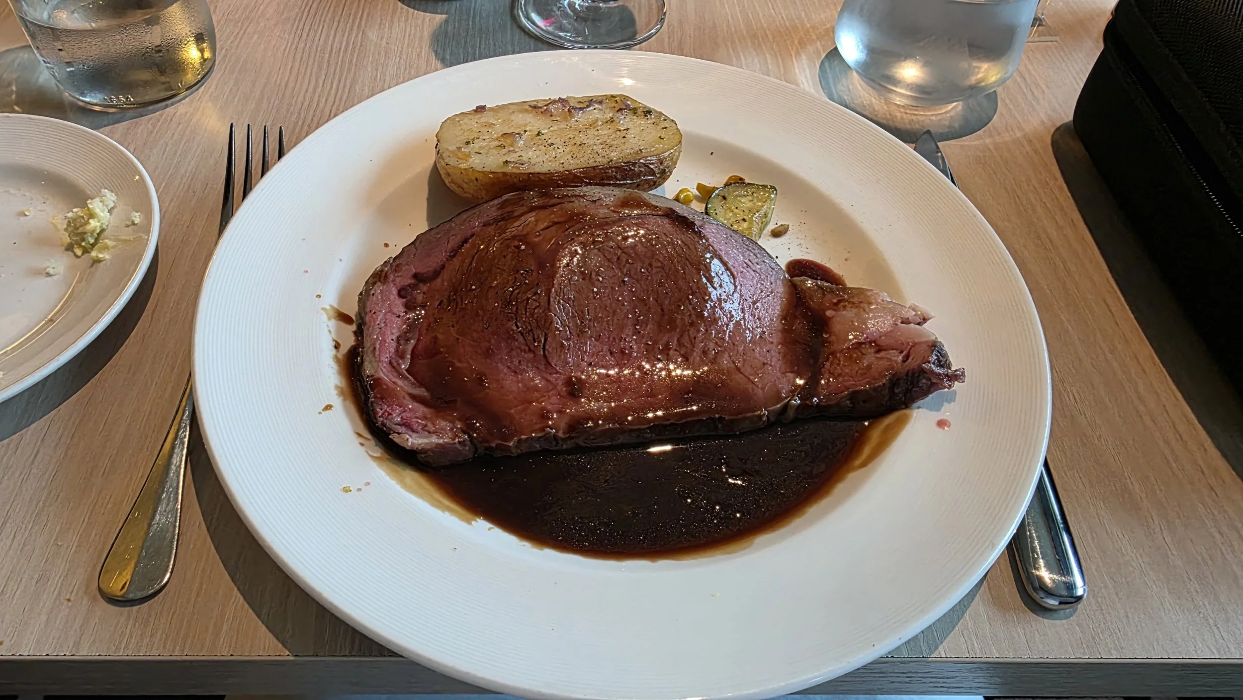A plate of cooked beef steak with sauce, a baked potato, and a small piece of zucchini, on a wooden table with cutlery and glasses nearby.