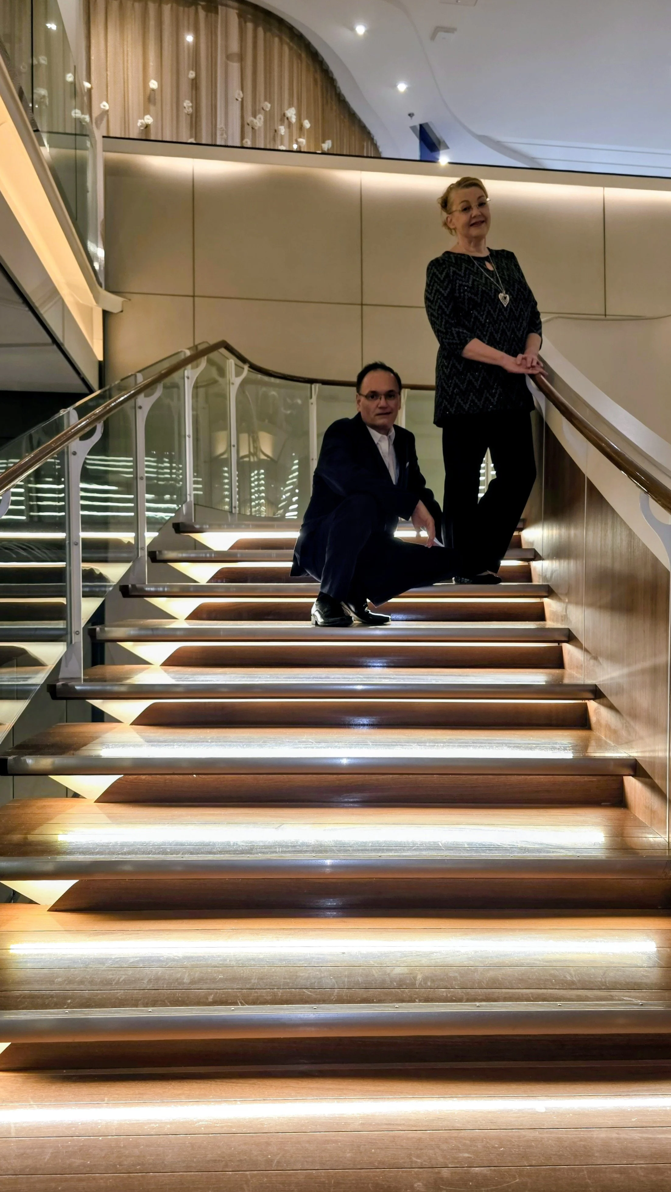 Two people, a woman and a man, posing on a spiral staircase in a modern indoor space with wood and glass decor.