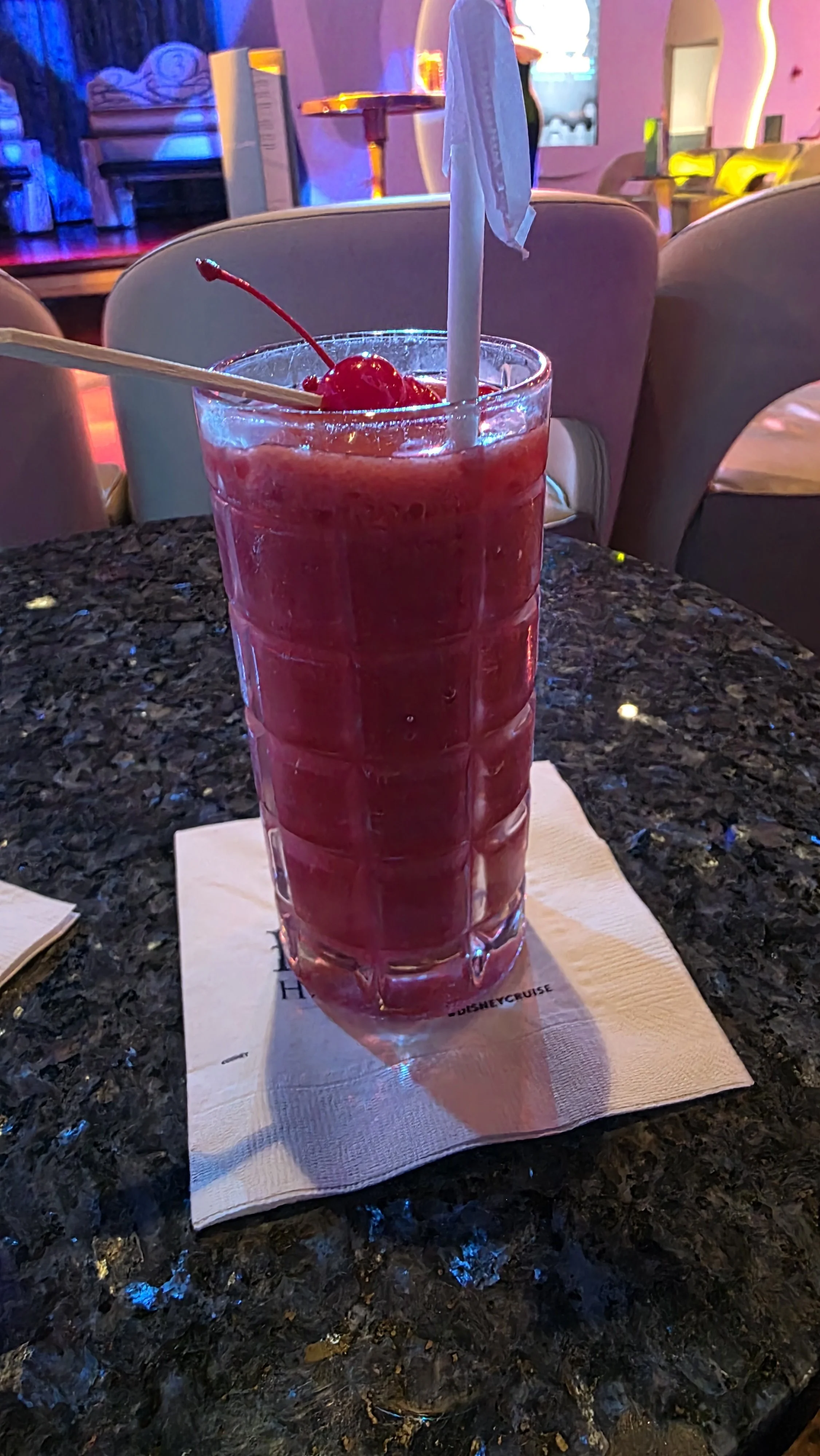 A tall glass filled with a pink, frozen cherry drink garnished with a maraschino cherry and a straw, placed on a napkin on a dark, polished table in a lively restaurant or bar setting.