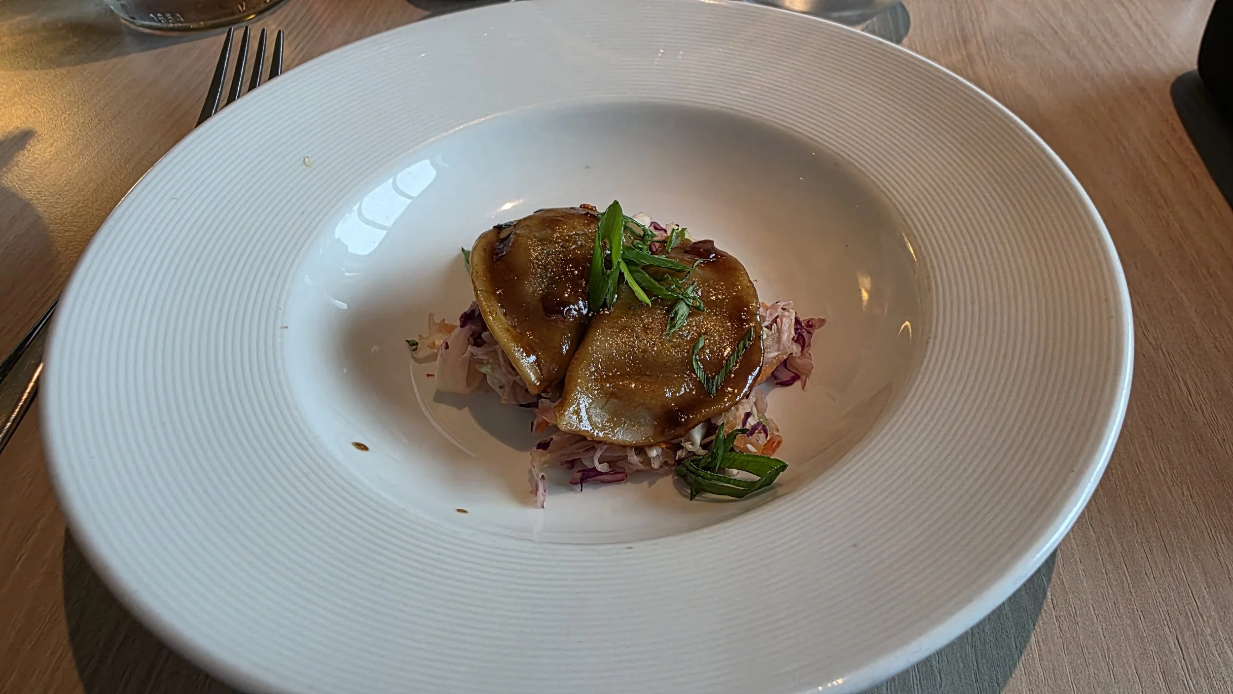 A white plate with a textured rim holding two pieces of glazed cooked meat garnished with green herbs, atop a bed of shredded vegetables, on a wooden table with silverware nearby.