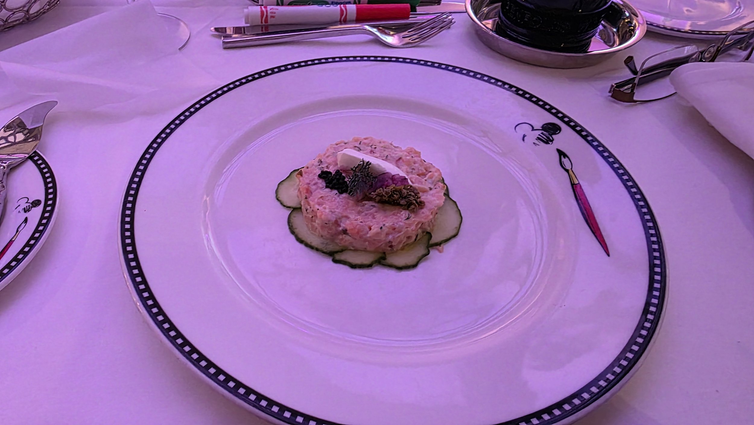 A fancy plate with an elegant, pink-colored seafood dish garnished with herbs, placed on a bed of sliced cucumbers. The plate has a unique decorative border design, resembling a movie reel or film strip.