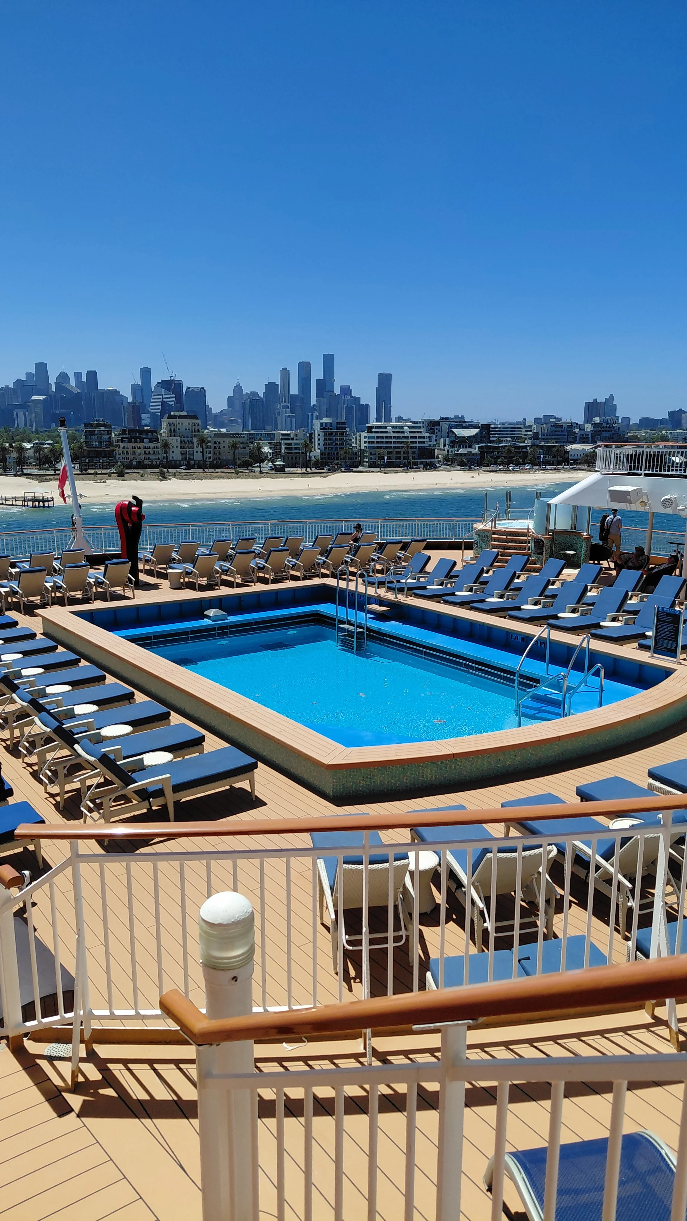 Pool area on a cruise ship deck with lounge chairs, overlooking a body of water and a city skyline in the background.