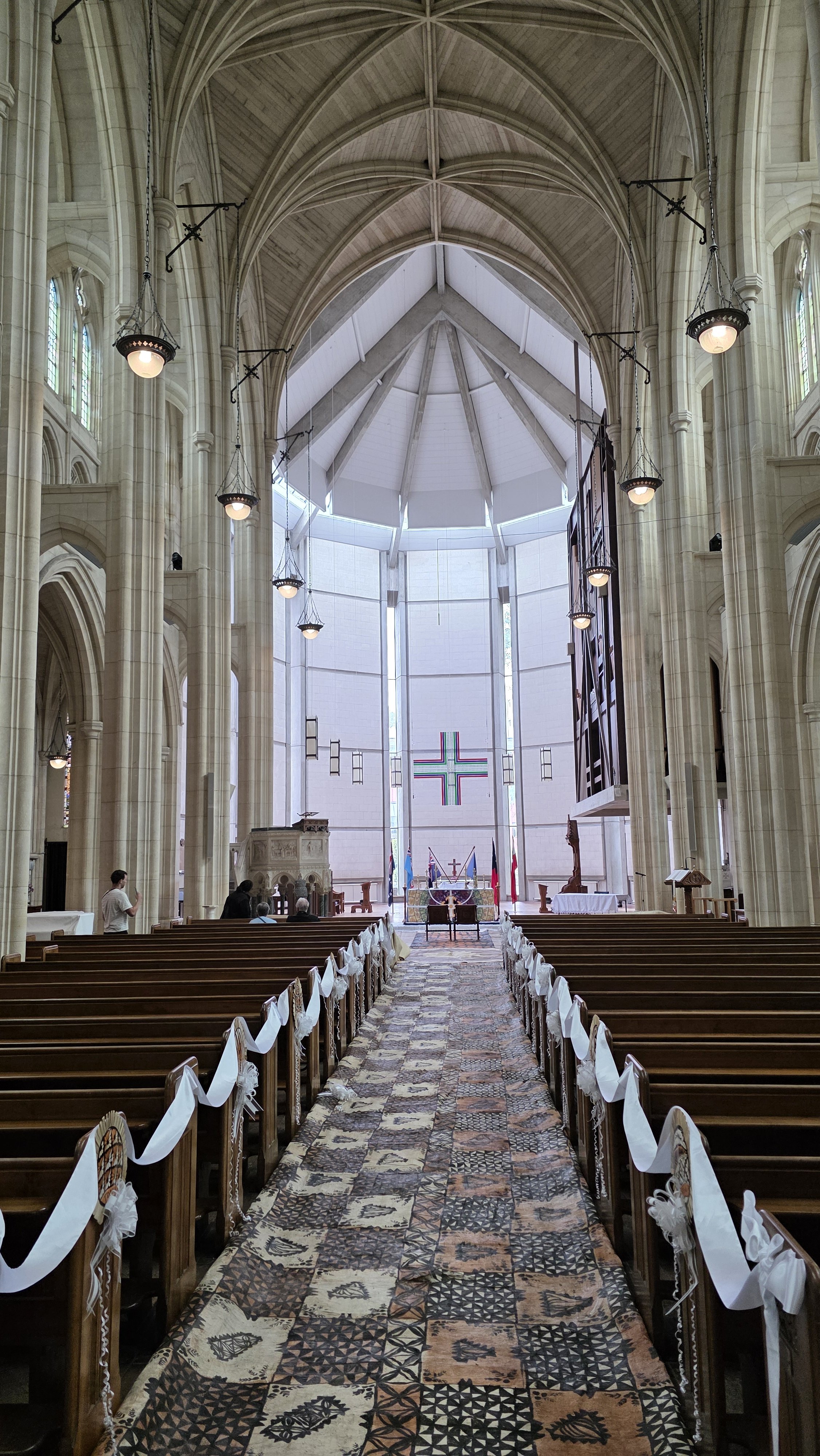Interior of a church with wooden pews decorated with white ribbons, a colorful carpeted aisle, and stained glass windows, leading to an altar with flags and a large cross.