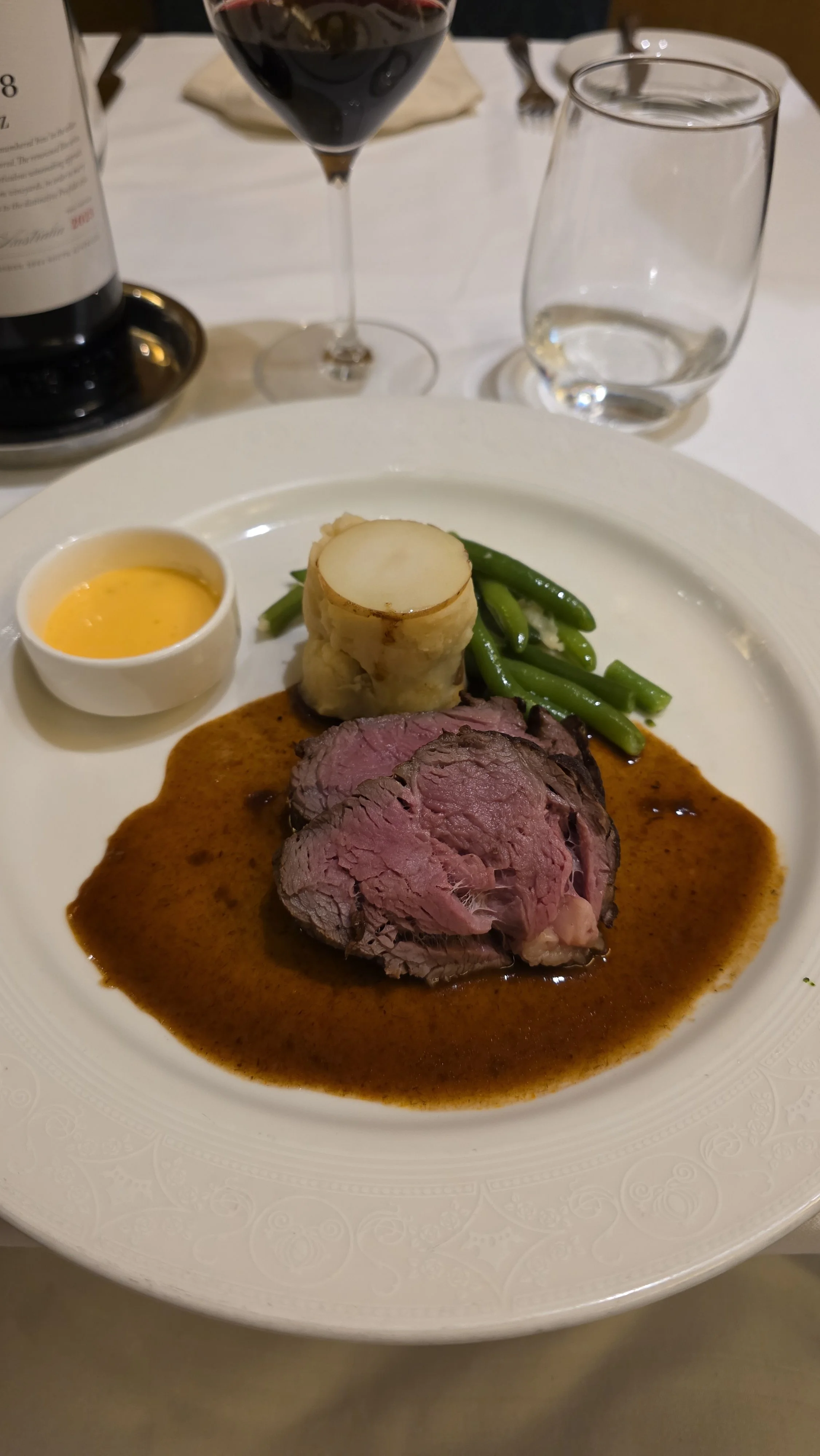 A plate of prime rib with green beans, a baked potato topped with sour cream, and a small dish of horseradish sauce, served with a glass of red wine and a glass of water.