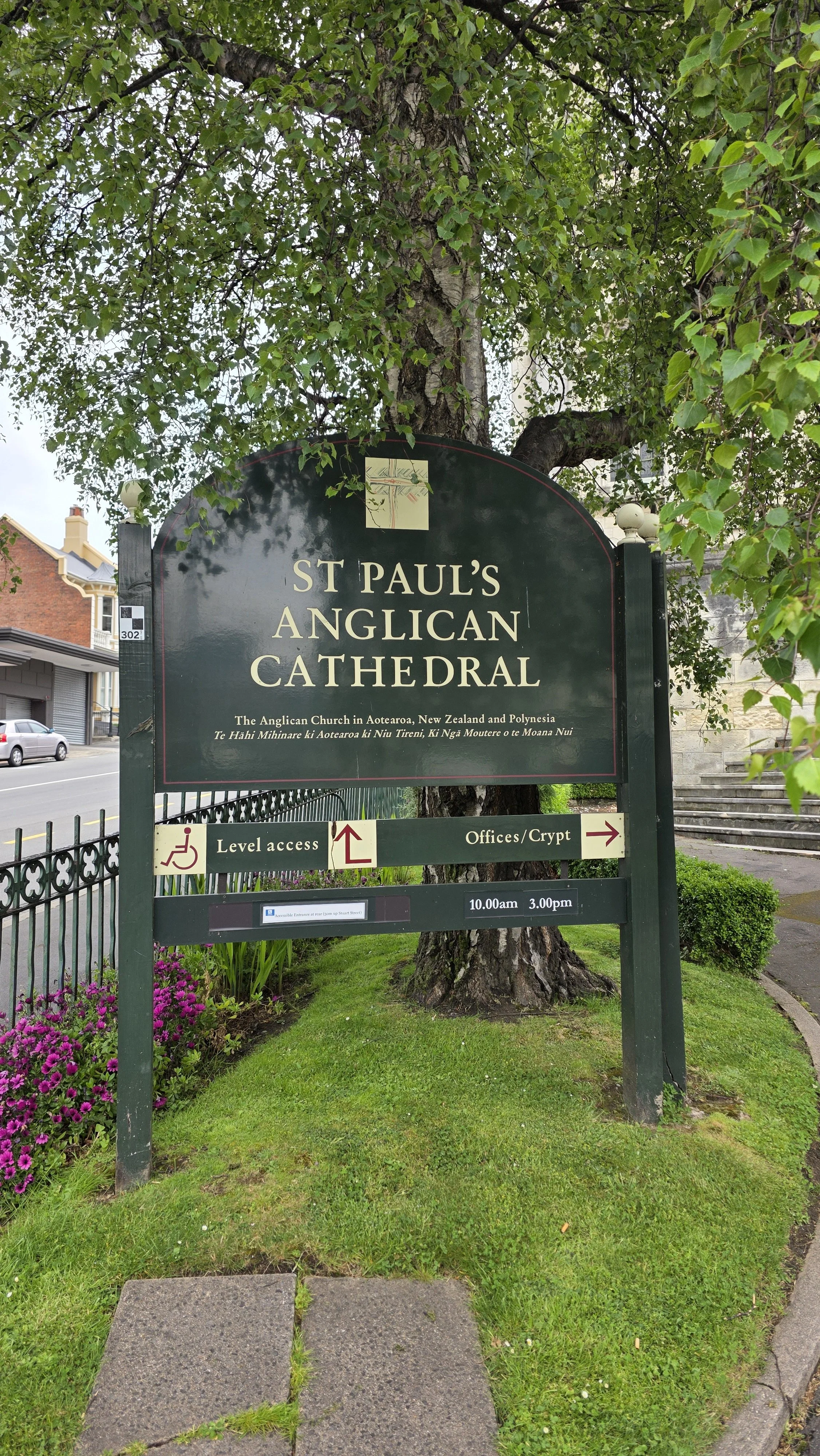 Sign for St. Paul's Anglican Cathedral in New Zealand. The sign provides directions for level access and offices/crypt, with opening hours from 10:00 am to 3:00 pm. The background features a large leafy tree and a building with stairs.