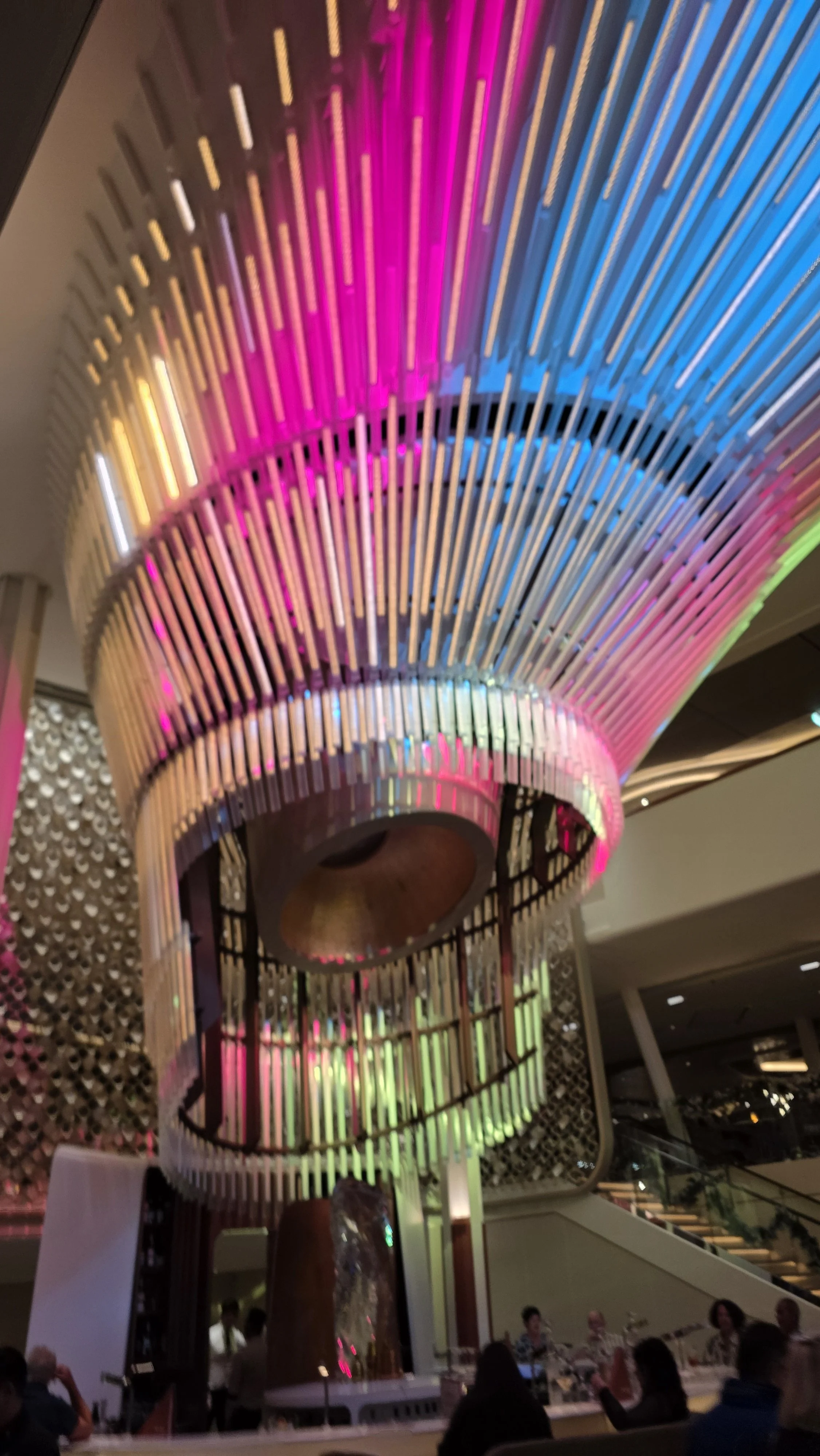 Colorful illuminated spiral chandelier hanging over a dining area with people seated at tables.