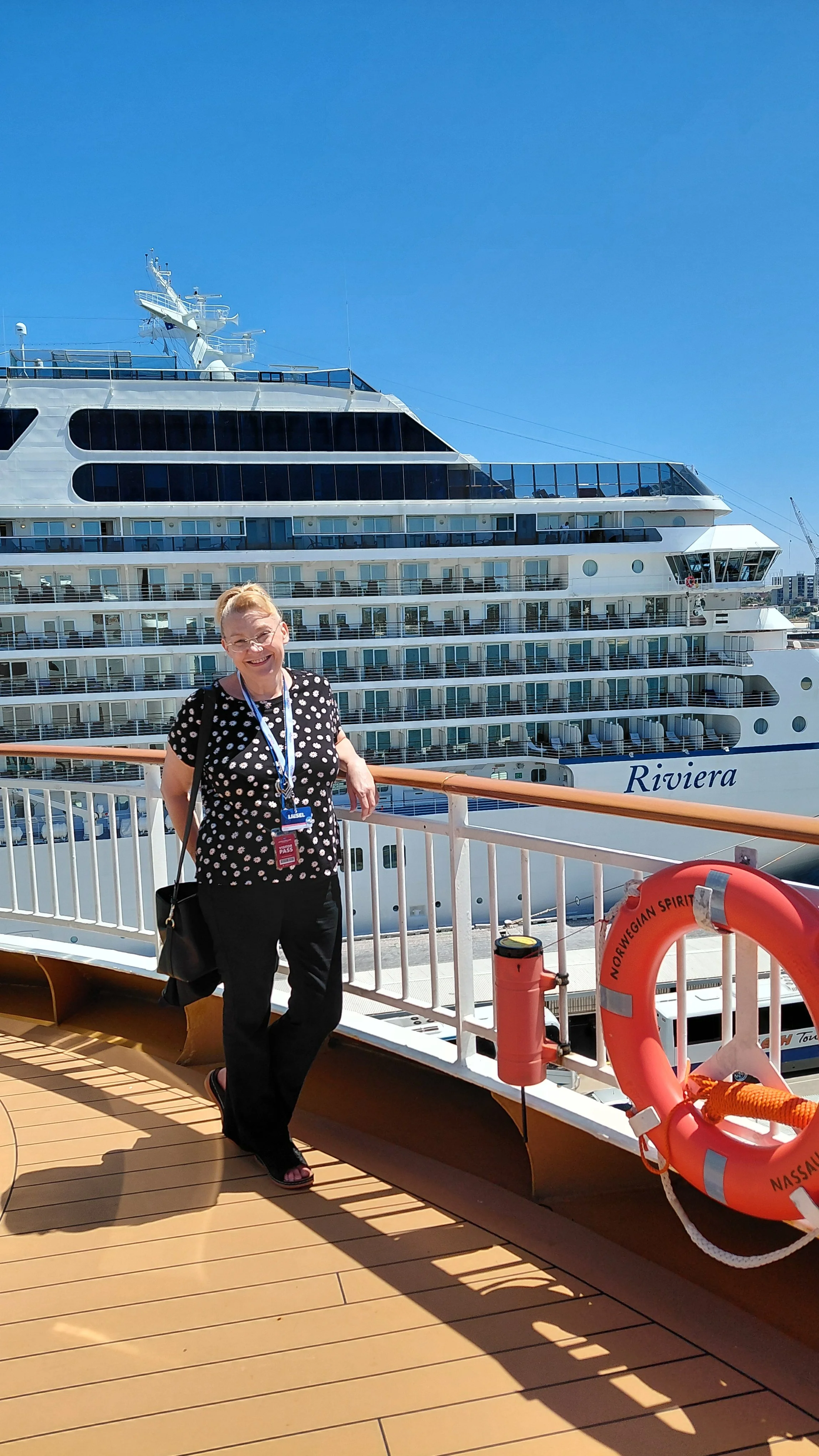 A smiling woman standing on a cruise ship deck with a large cruise ship named Riviera in the background, clear blue sky overhead, and safety equipment nearby.
