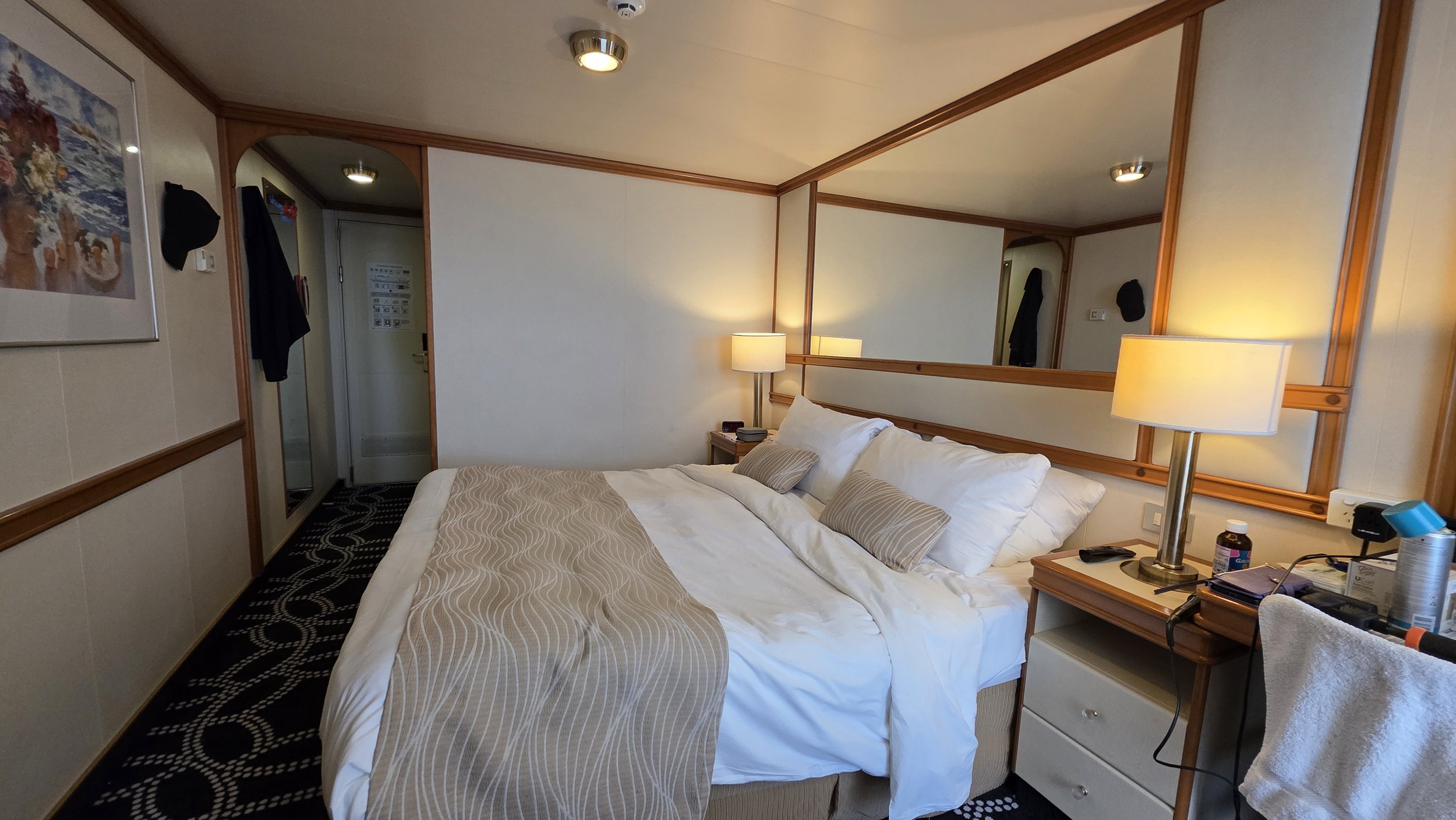A hotel room with a neatly made bed, bedside lamps, a mirror, artwork on the wall, and various personal items on the nightstand and table, with a doorway visible in the background.