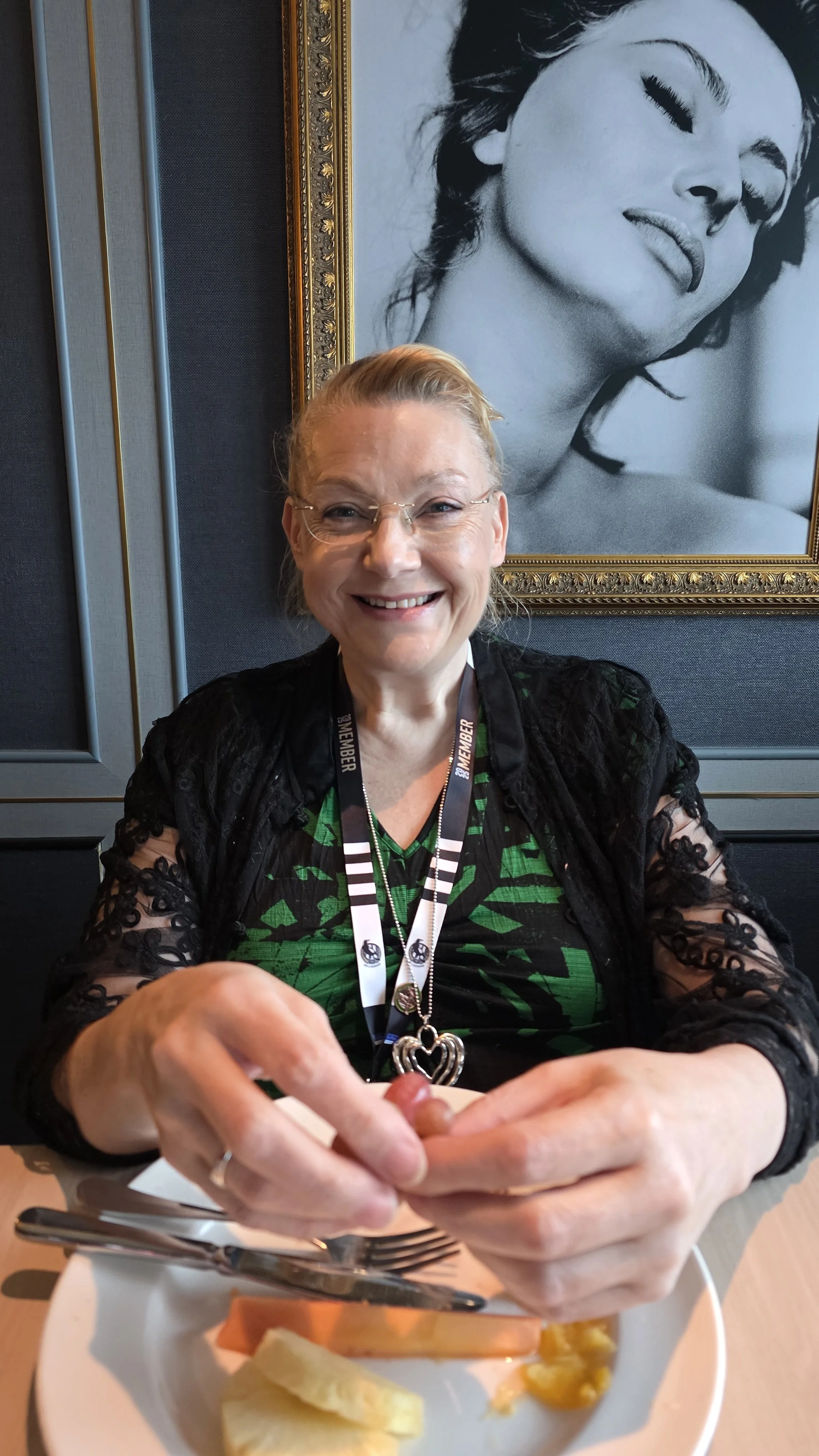 A smiling woman with blonde hair and glasses sitting at a table with food, holding a strawberry, in a restaurant with a large black-and-white portrait of a woman in the background.