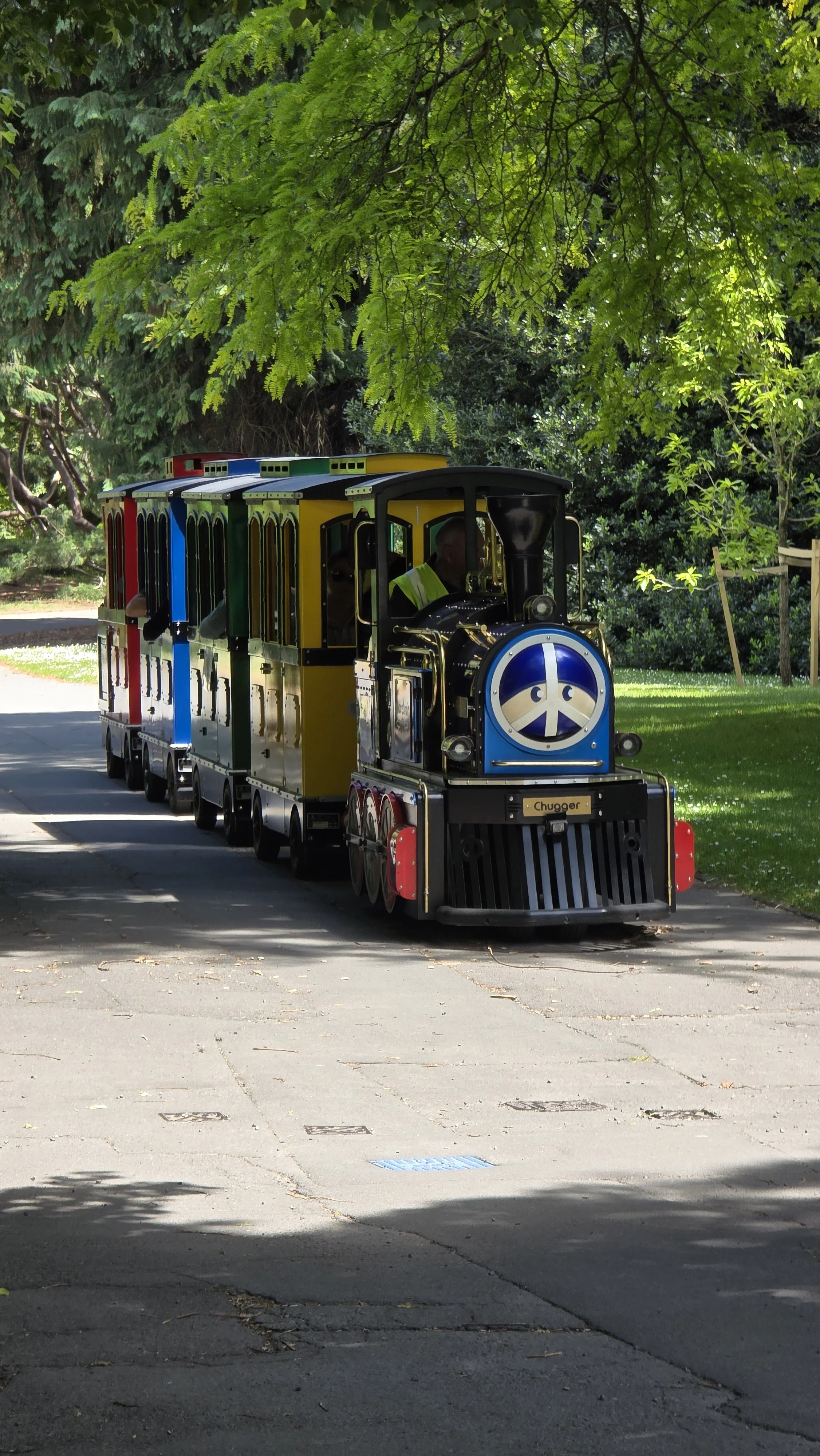 Colorful small train with a peace sign on the front, traveling on a paved path in a park shaded by trees.