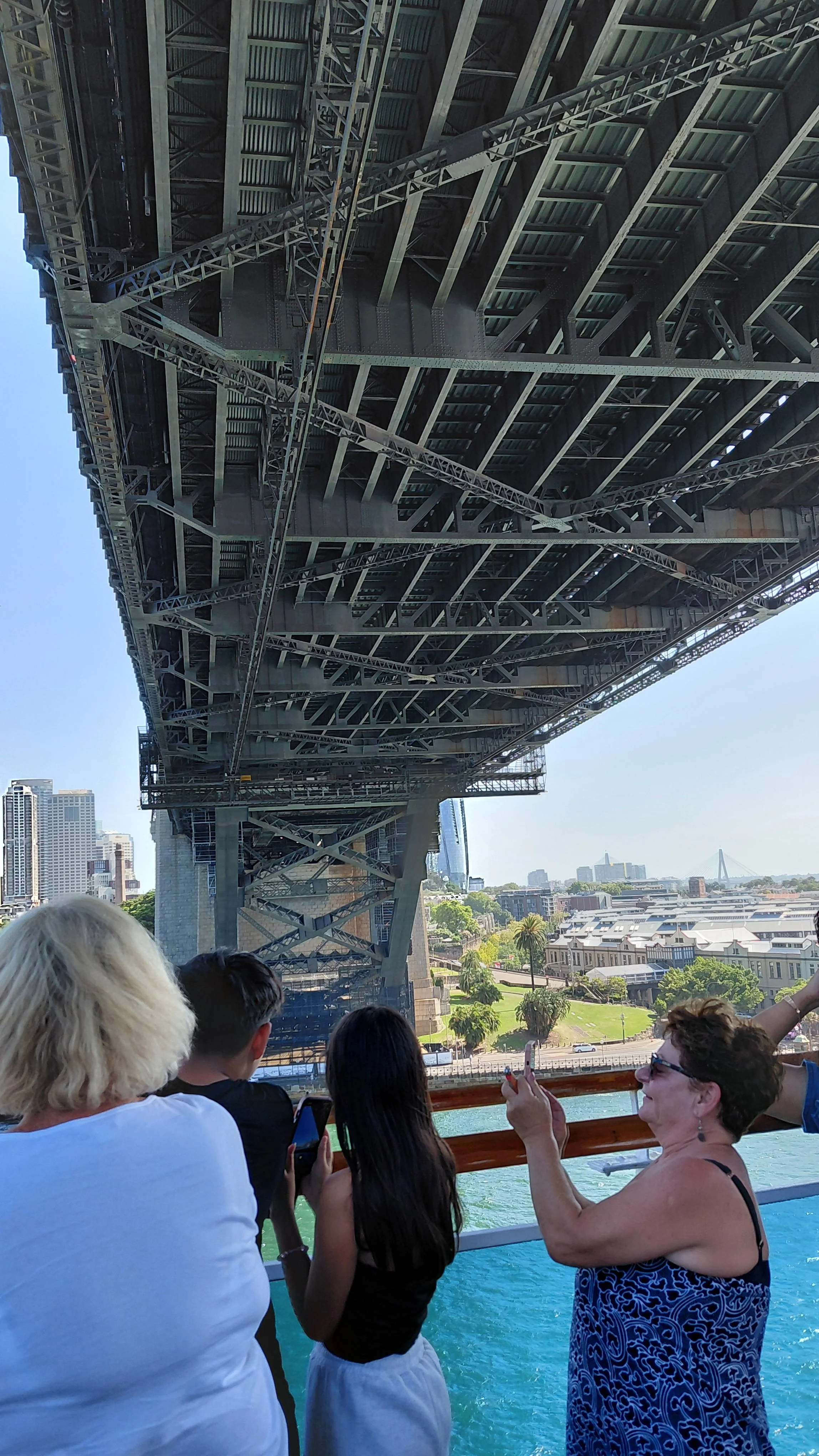 Tourists taking photos on a boat with a view of the underside of a large bridge and cityscape in the background.